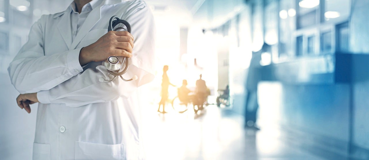 Doctor standing in the middle of hospital hallway wearing a white long lab coat with arms crossed and stethoscope in hand, with patients in the back.