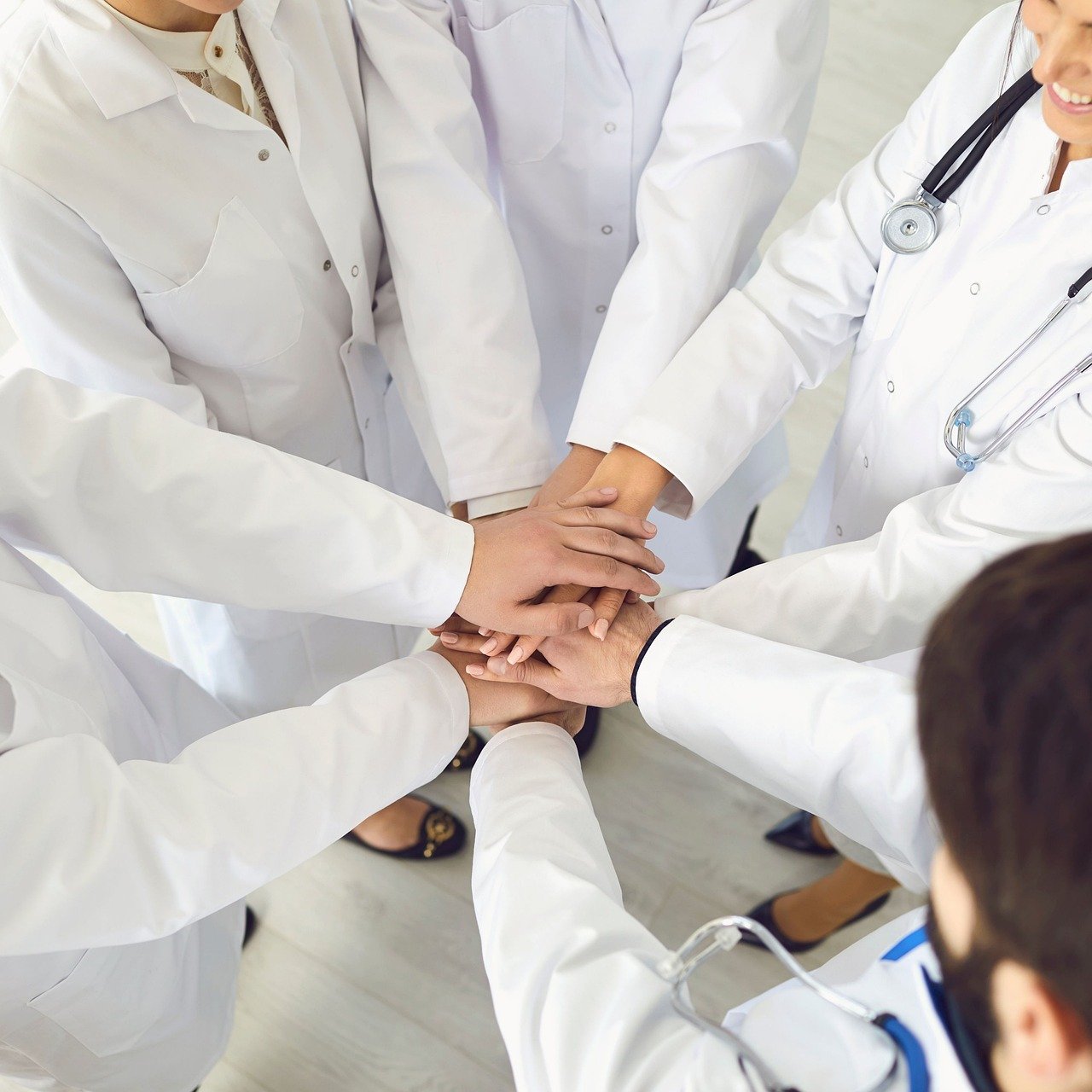 Five doctors and nurses wearing white lab coats and stethoscopes putting their clean hands together before starting their morning rounds.