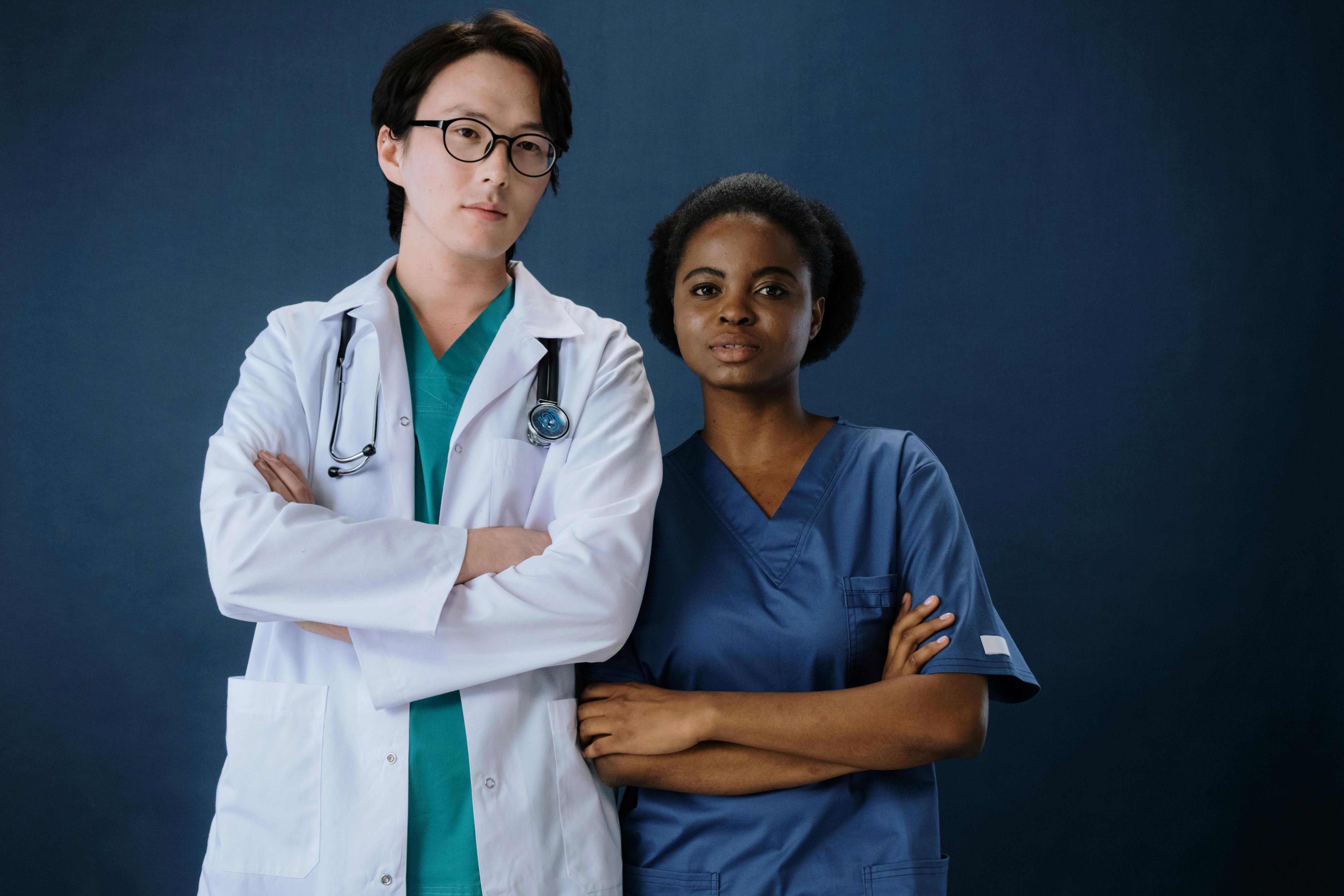 A doctor wearing a white lab coat, teal scrub top, teal scrub pants and a traveling nurse wearing an admiral blue scrub top and scrub pants. 