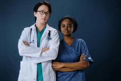 Image of A doctor wearing a white lab coat, teal scrub top, teal scrub pants and a traveling nurse wearing an admiral blue scrub top and scrub pants. 