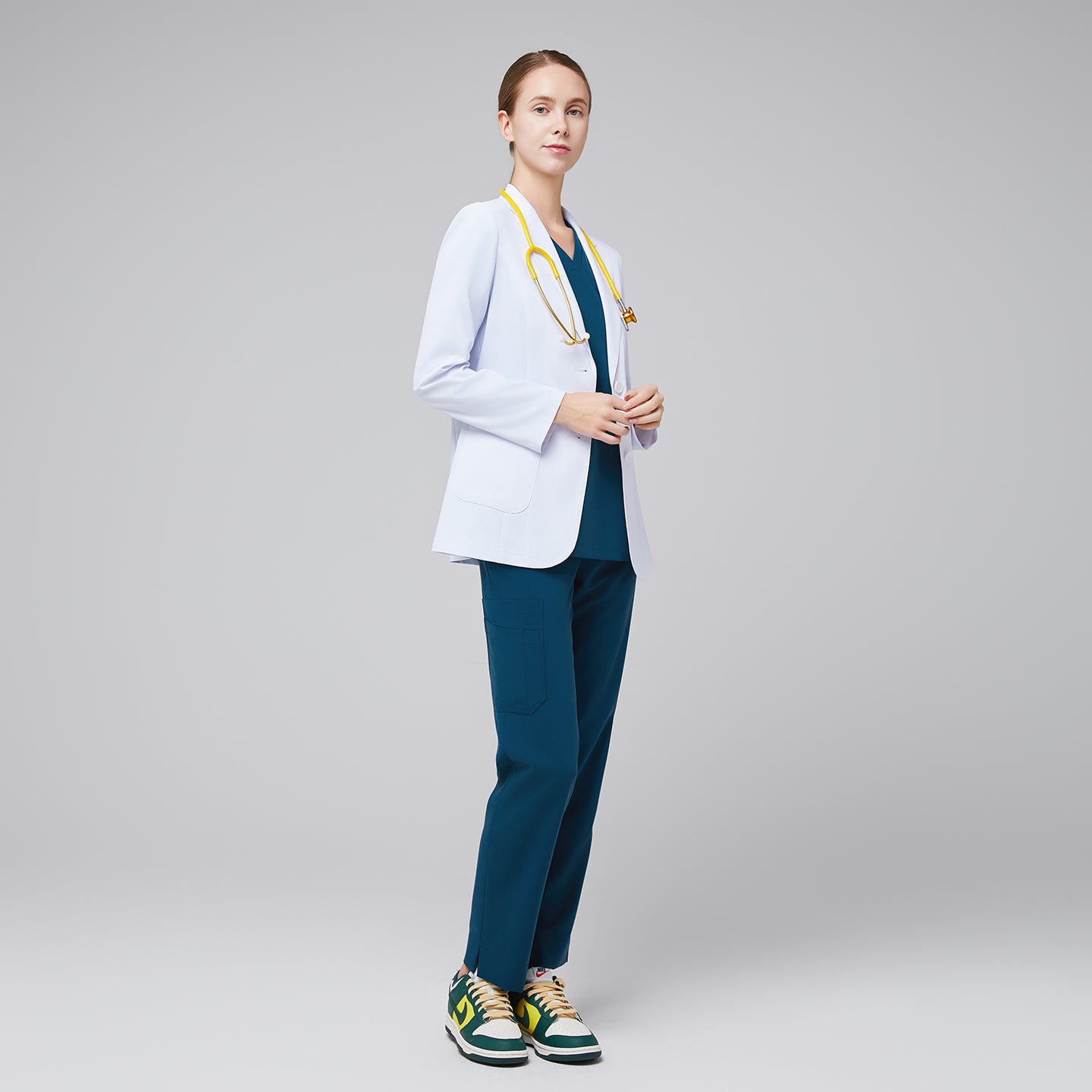 Female medical professional in a white lab coat over teal scrubs, with a yellow stethoscope around her neck, standing confidently with hands clasped,White