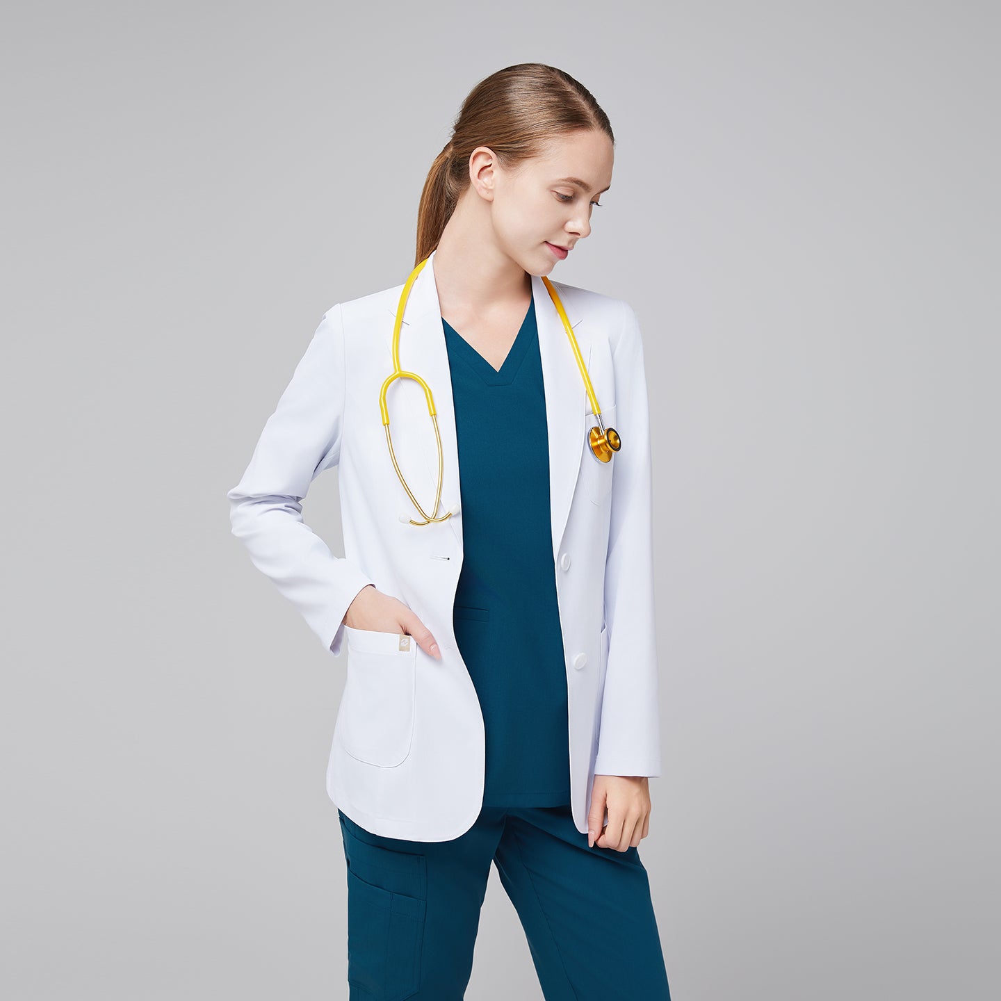 Female medical professional in a white lab coat over teal scrubs, wearing a yellow stethoscope, with one hand in her pocket, looking down,White