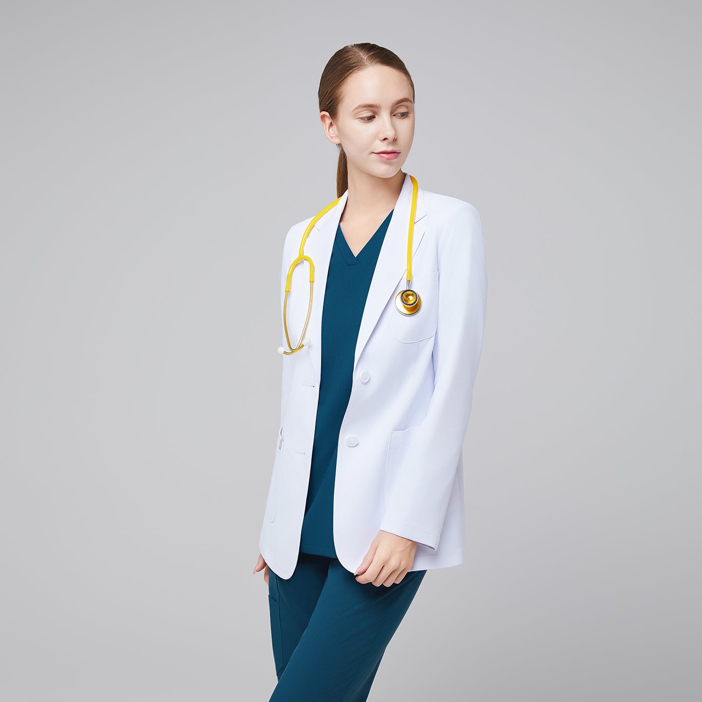 Female medical professional in a white lab coat over teal scrubs, wearing a yellow stethoscope around her neck, looking to the side,White