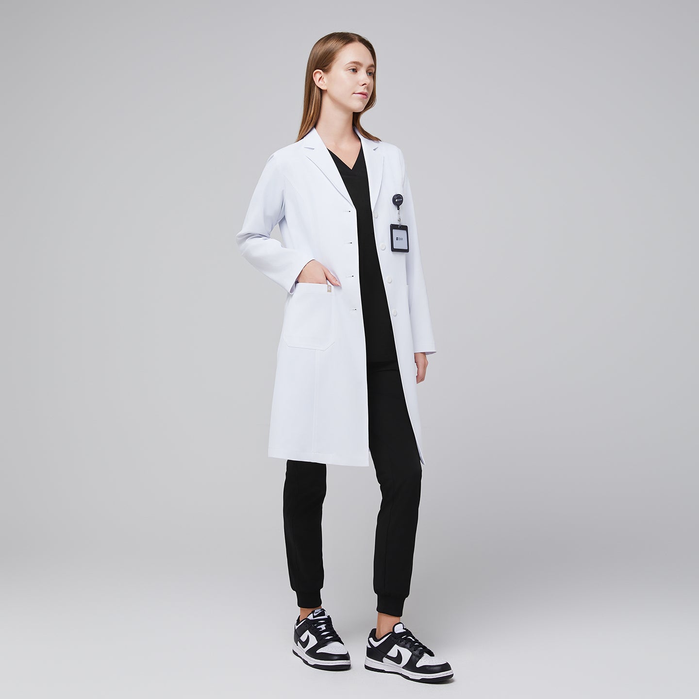 Female doctor in white lab coat and black scrubs with ID badge, standing confidently,White