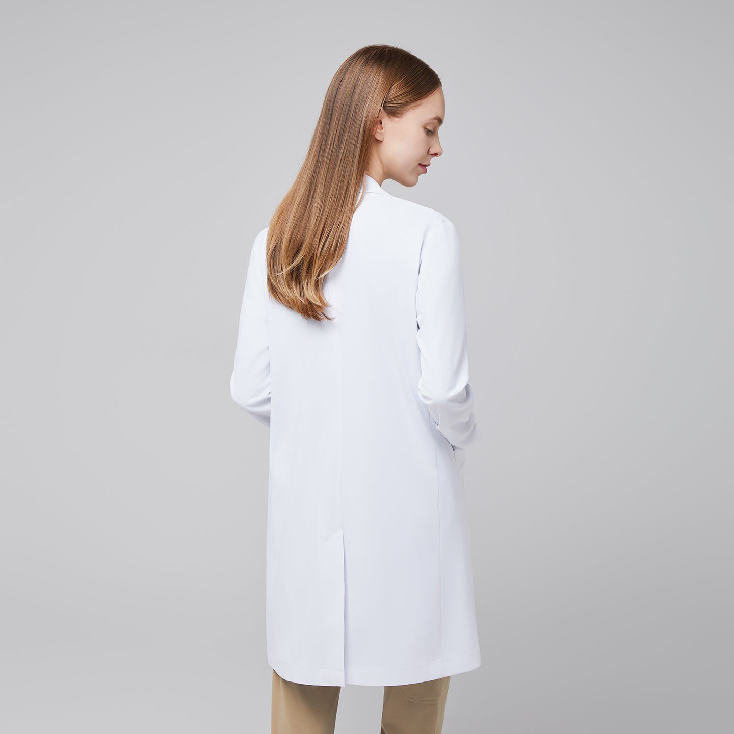 Model with her back turned, wearing a white long lab coat with chest and side pockets, with a sand beige scrub top and bottom under,White