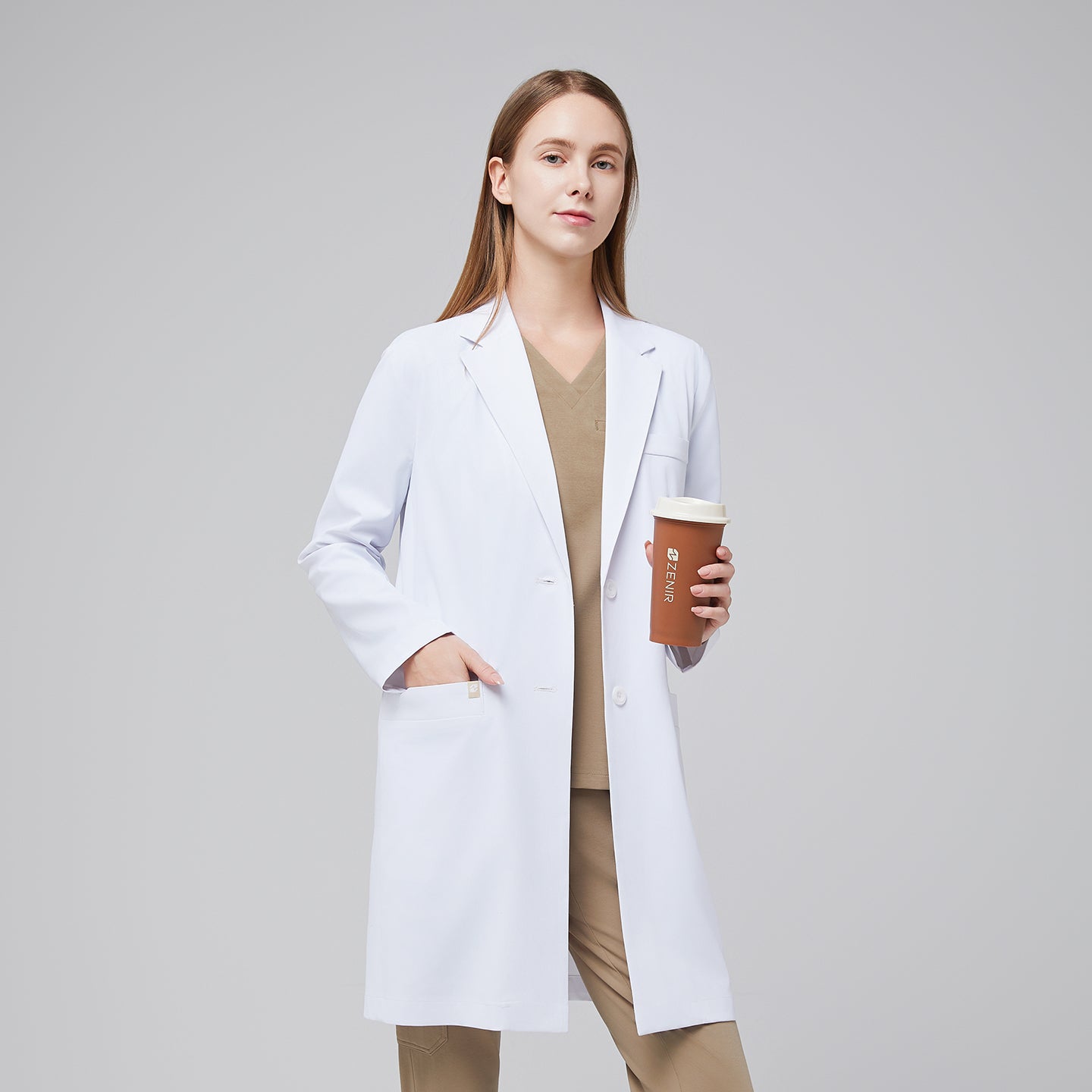 Model facing forward with a hand in her pocket, wearing a white long lab coat with chest and side pockets, with a sand beige scrub top and bottom under, holding a brown tumbler,White