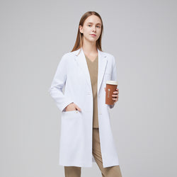 Image of Model facing forward with a hand in her pocket, wearing a white long lab coat with chest and side pockets, with a sand beige scrub top and bottom under, holding a brown tumbler,White