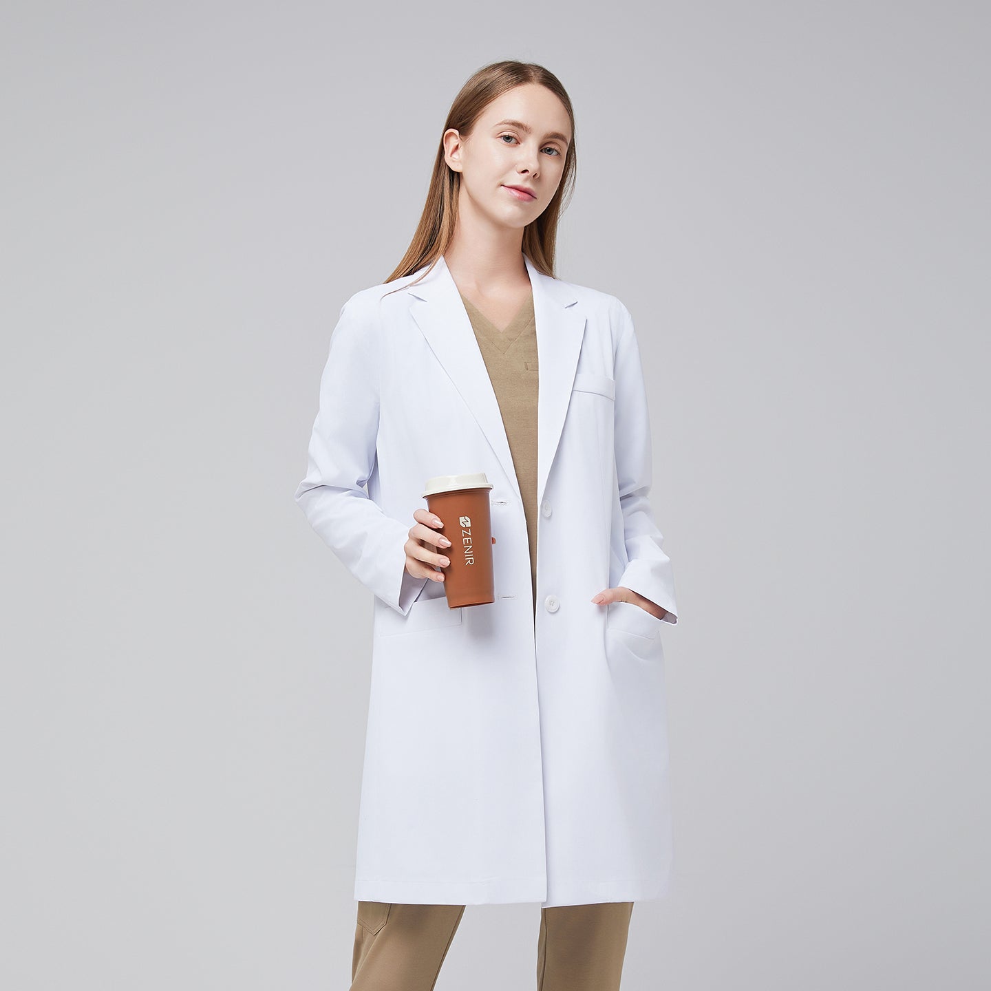 Smiling model wearing a white long lab coat with chest and side pockets, with a sand beige scrub top and bottom under, holding a brown tumbler,White