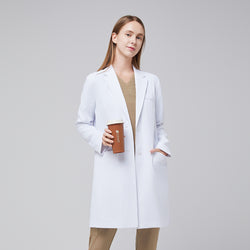 Image of Smiling model wearing a white long lab coat with chest and side pockets, with a sand beige scrub top and bottom under, holding a brown tumbler,White