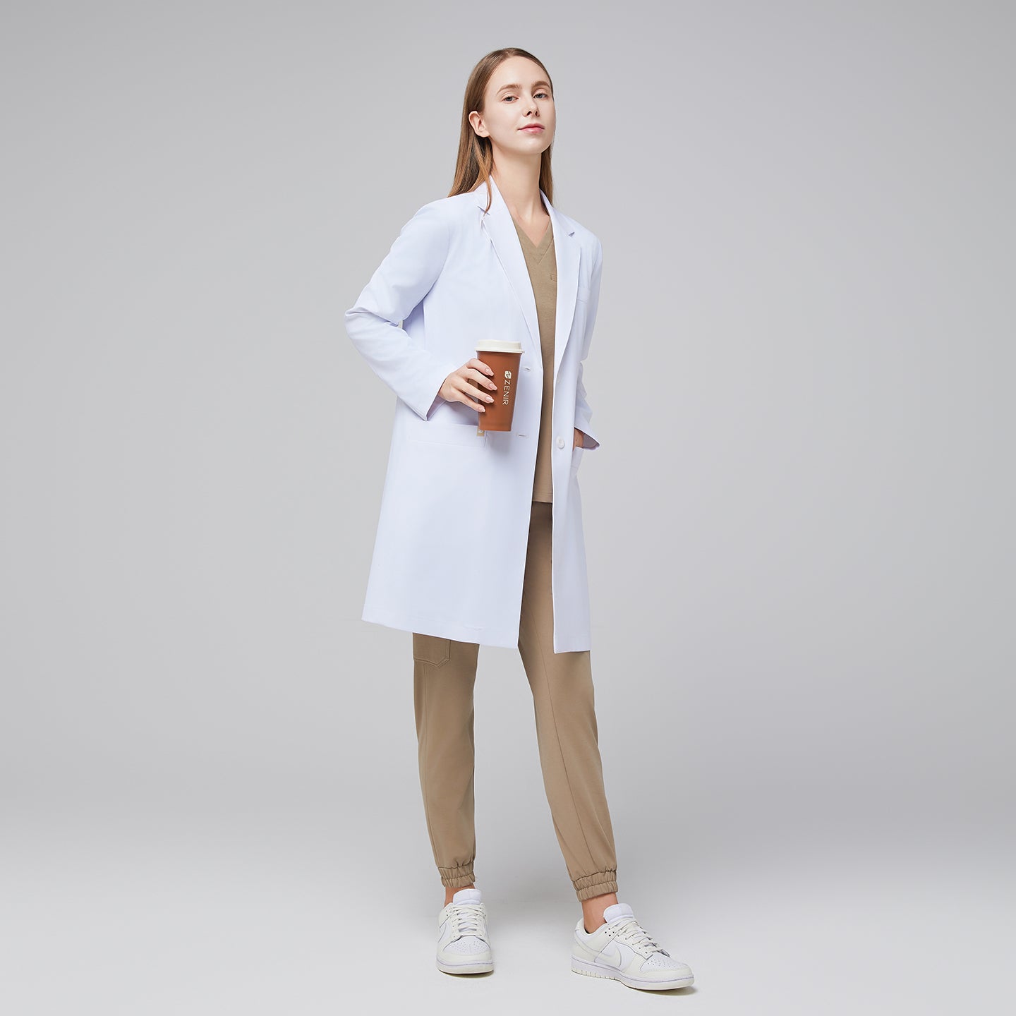 Model facing forward with a hand in her pocket, wearing a white long lab coat with chest and side pockets, with a sand beige scrub top and bottom under, holding a brown tumbler,White