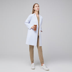 Image of Model facing forward with a hand in her pocket, wearing a white long lab coat with chest and side pockets, with a sand beige scrub top and bottom under, holding a brown tumbler,White