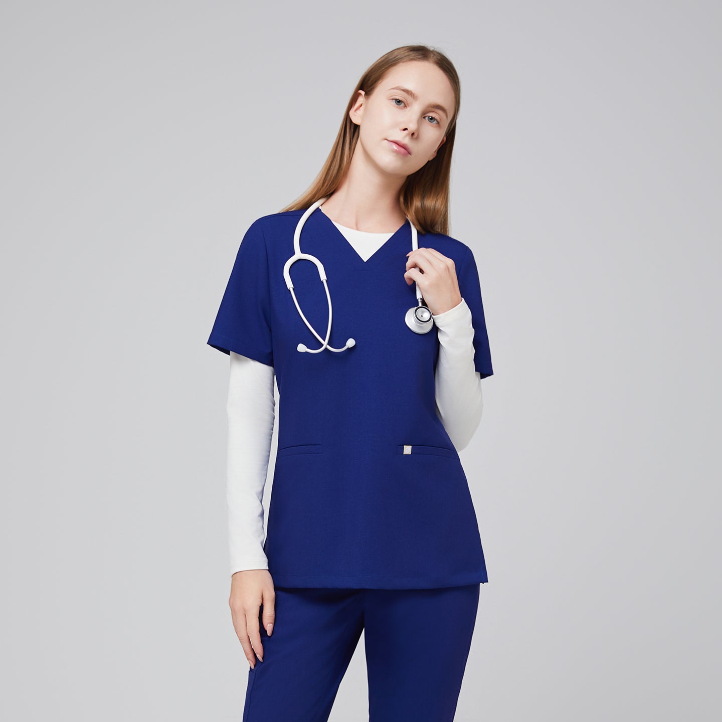 Model wearing a navy blue double pocket V-neck scrub top layered over a white long sleeve shirt, with a stethoscope around the neck,Admiral Blue