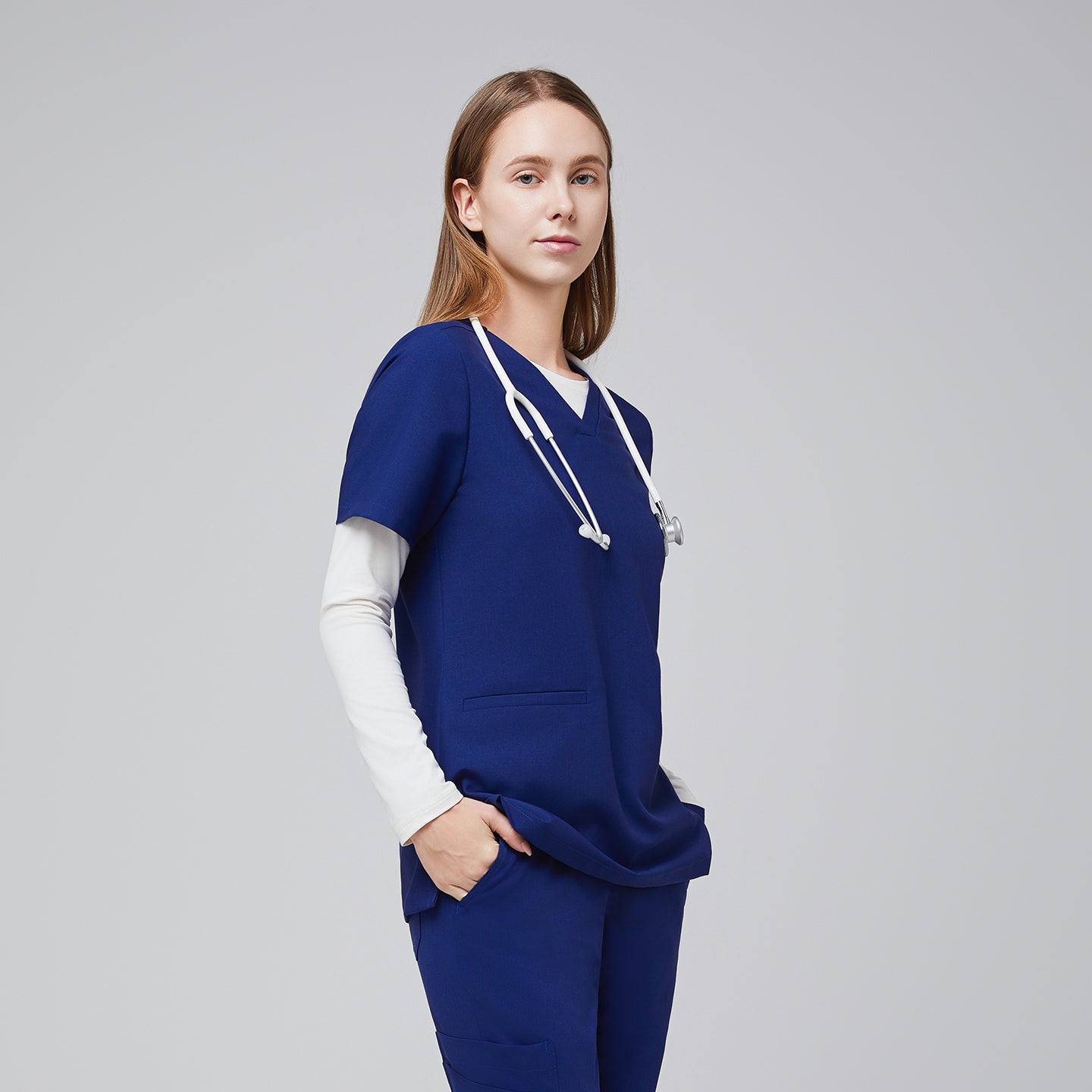 Model in a navy blue double pocket V-neck scrub top, layered over a white long sleeve shirt, with a stethoscope, and matching scrub pants,Admiral Blue