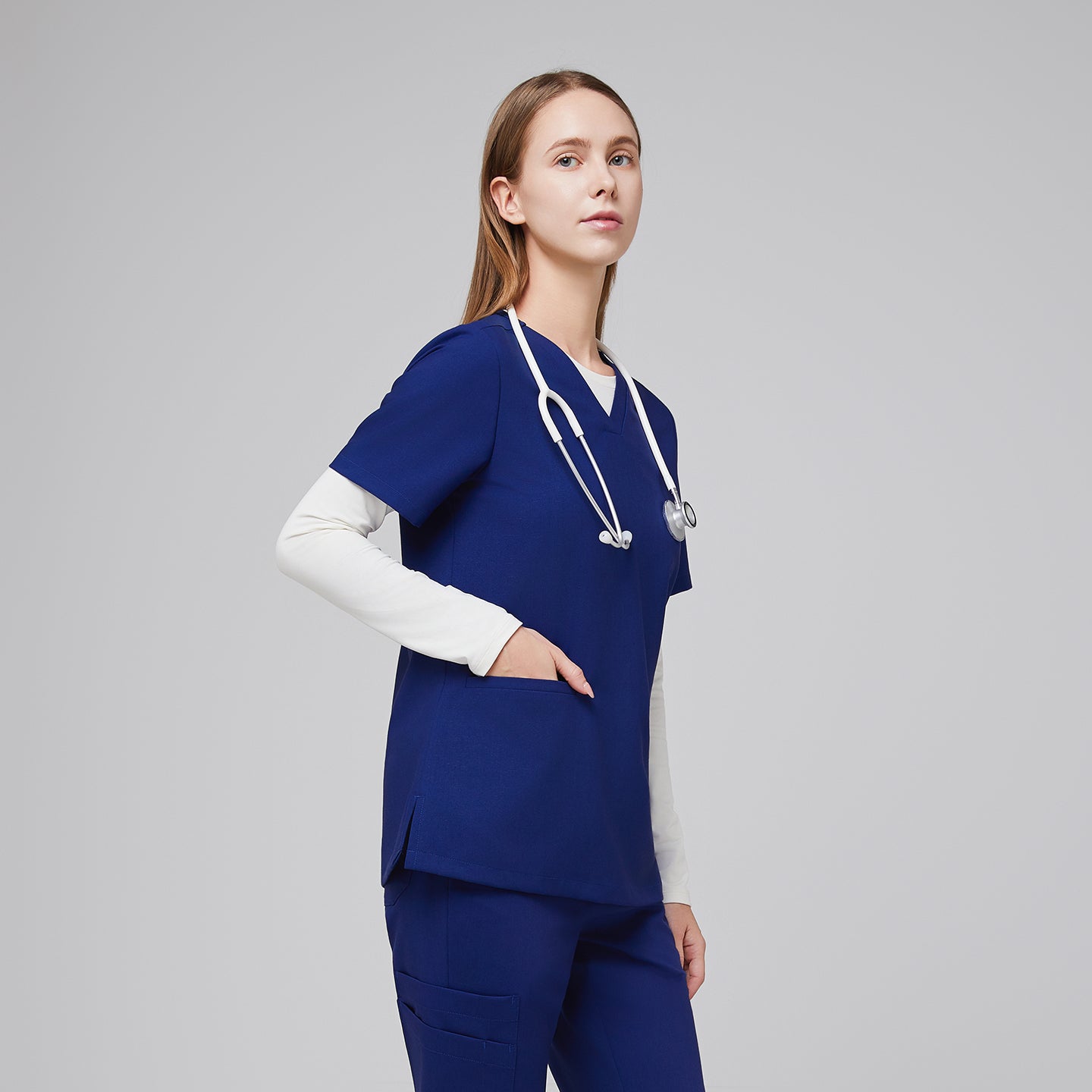 Model wearing a navy blue double pocket V-neck scrub top over a white long sleeve shirt, holding a stethoscope, and matching scrub pants,Admiral Blue