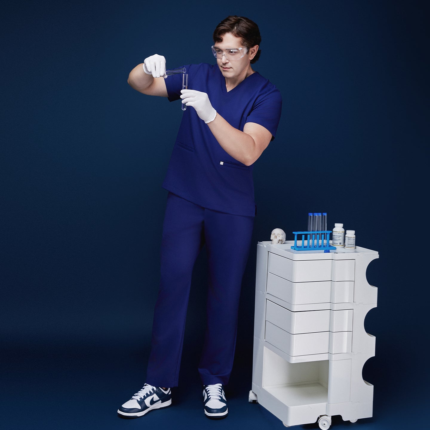 Man in admiral blue scrubs examining a test tube, wearing gloves and goggles, standing beside a medical cart,Admiral Blue