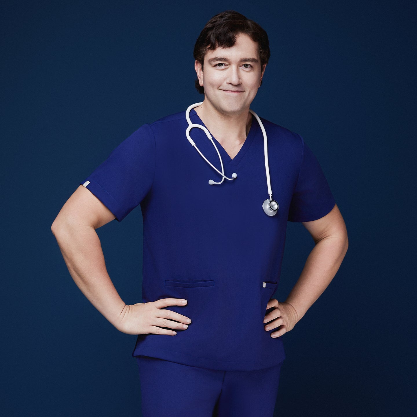 Man in admiral blue scrubs with V-neck and double pockets, wearing a white stethoscope, posing confidently with hands on hips and a smile,Admiral Blue