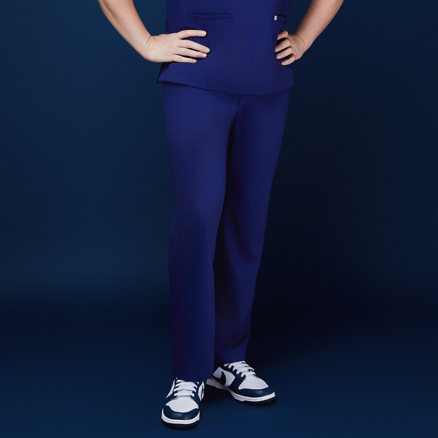 Front view of model wearing admiral blue scrub pants with a pocket design, paired with navy and white sneakers, against a navy backdrop,Admiral Blue