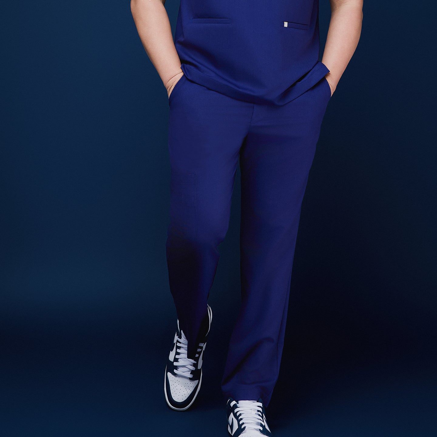 Model showcasing admiral blue scrub pants with hands in pockets, paired with sneakers, standing against a navy backdrop,Admiral Blue