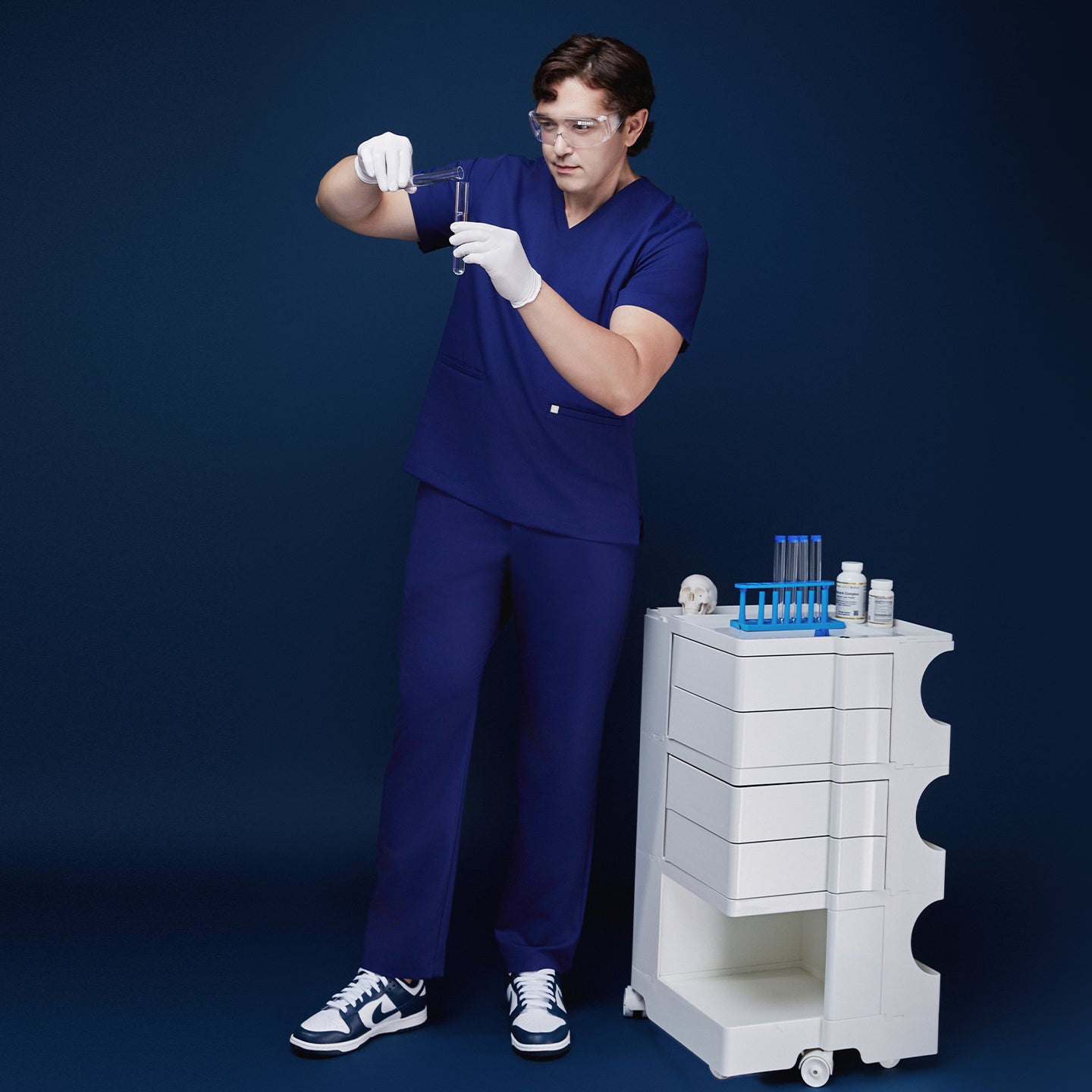 Model in admiral blue scrub set conducting a lab experiment, holding test tubes while wearing gloves and safety glasses, standing next to a medical cart with lab equipment, against a navy backdrop,Admiral Blue