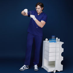 Image of Model in admiral blue scrub set conducting a lab experiment, holding test tubes while wearing gloves and safety glasses, standing next to a medical cart with lab equipment, against a navy backdrop,Admiral Blue