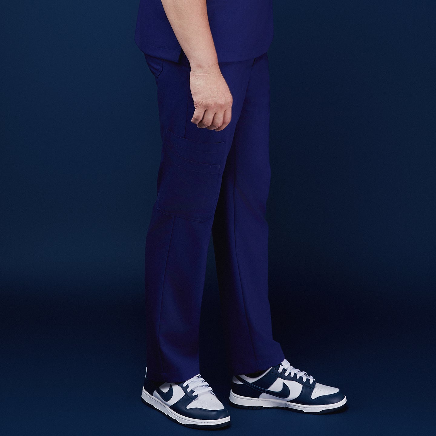 Side view of model wearing admiral blue scrub pants with a pocket design, paired with navy and white sneakers, against a navy backdrop,Admiral Blue