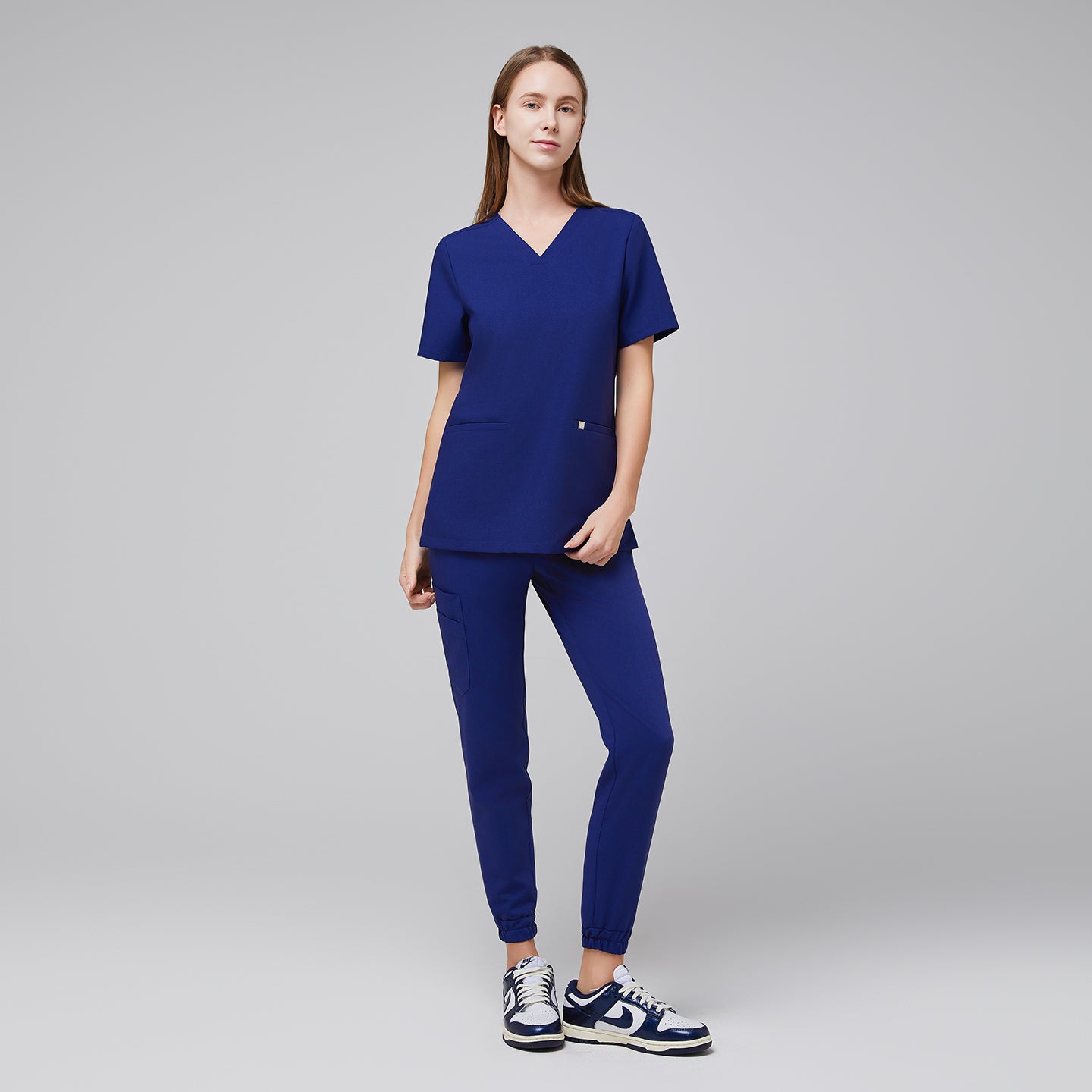 Model wearing admiral blue jogger scrub pants with elastic cuffs and a matching V-neck scrub top, paired with white and blue Nike sneakers,Admiral Blue