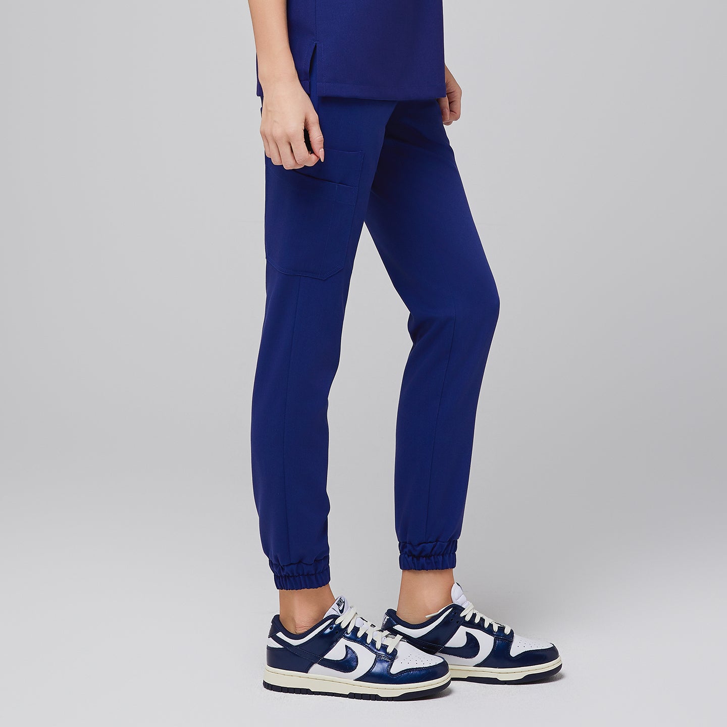 Side view of admiral blue jogger scrub pants with cargo pocket detail and elastic cuffs, paired with white and blue Nike sneakers,Admiral Blue