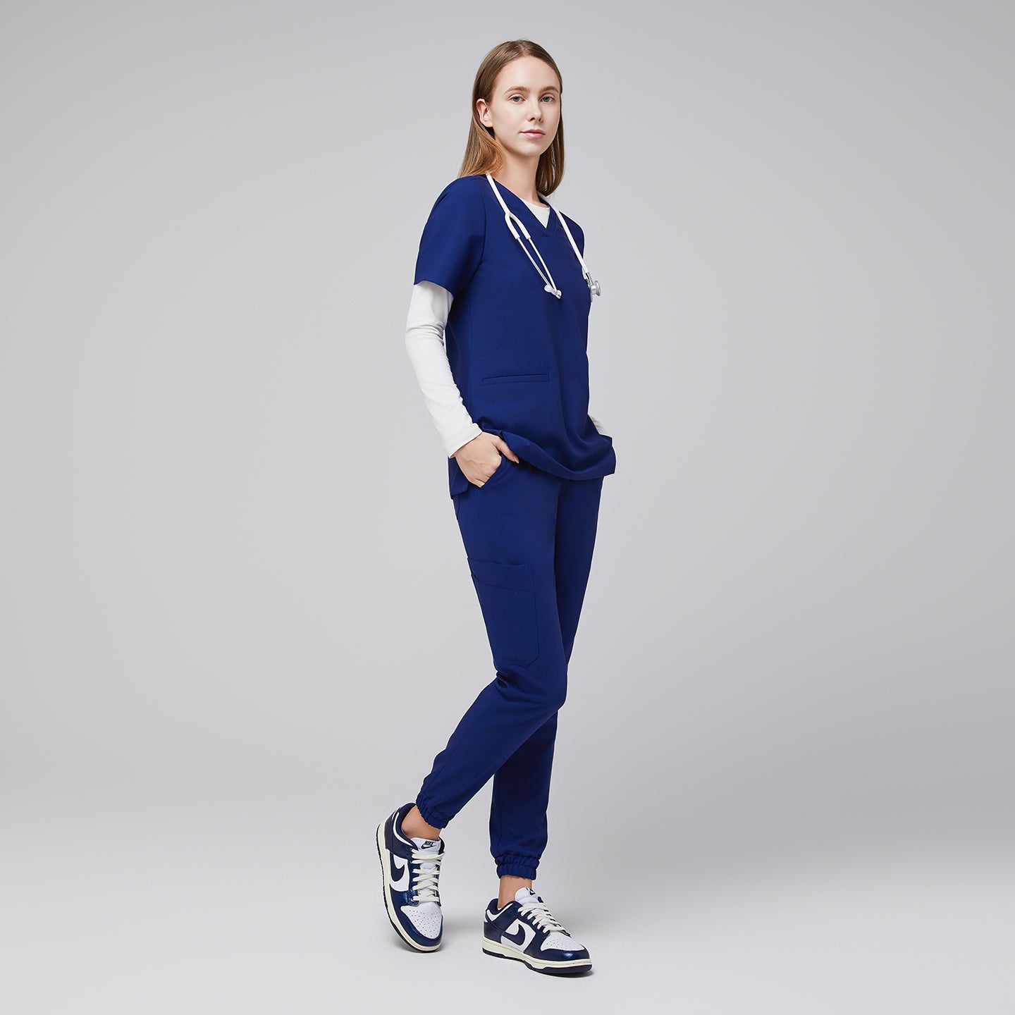 Model wearing admiral blue jogger scrub pants with elastic cuffs and a matching V-neck top, styled with a stethoscope and white and blue Nike sneakers,Admiral Blue