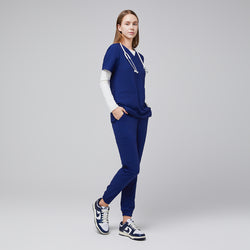 Image of Model wearing admiral blue jogger scrub pants with elastic cuffs and a matching V-neck top, styled with a stethoscope and white and blue Nike sneakers,Admiral Blue