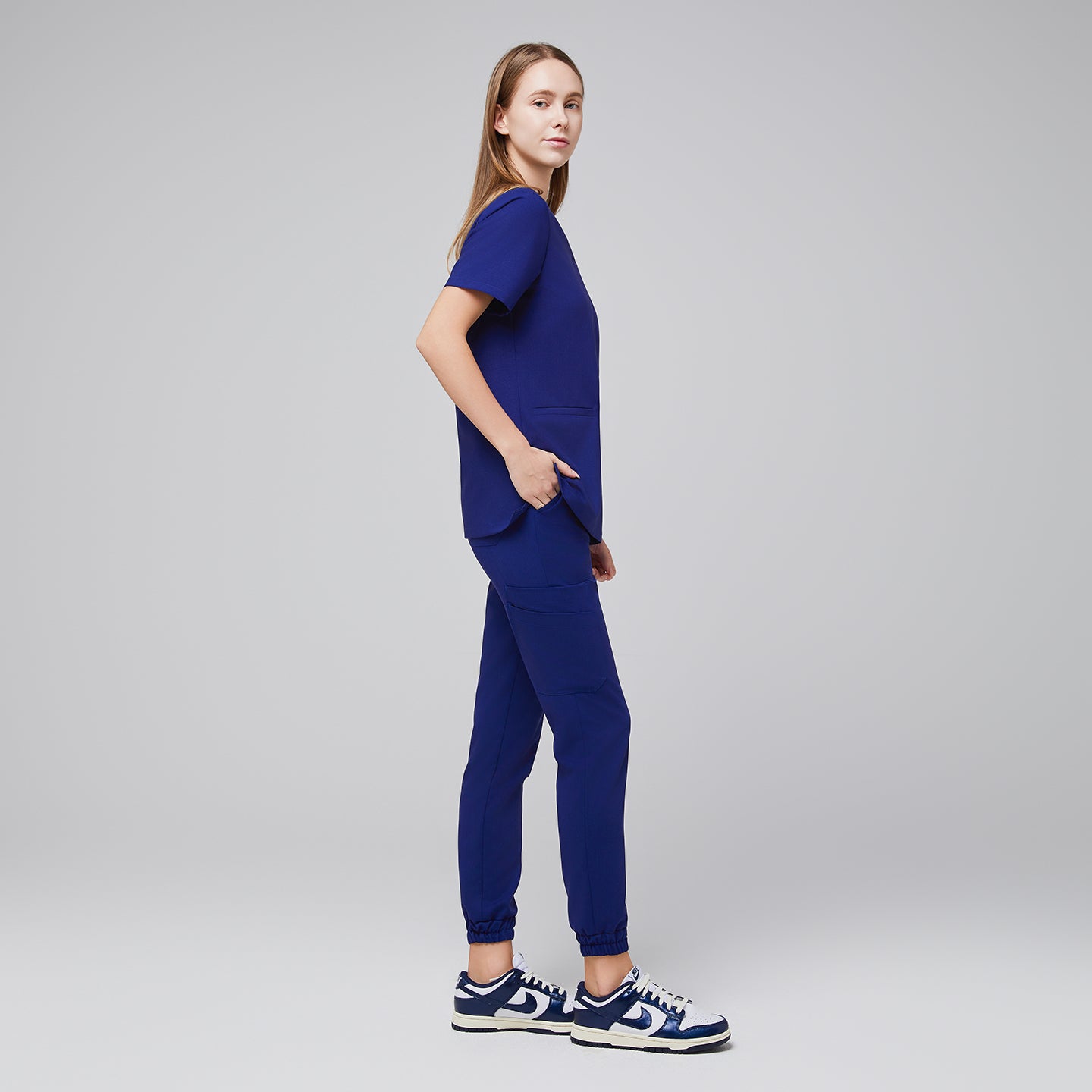 Side view of model wearing admiral blue jogger scrub pants with elastic cuffs and a matching V-neck top, paired with white and blue Nike sneakers,Admiral Blue