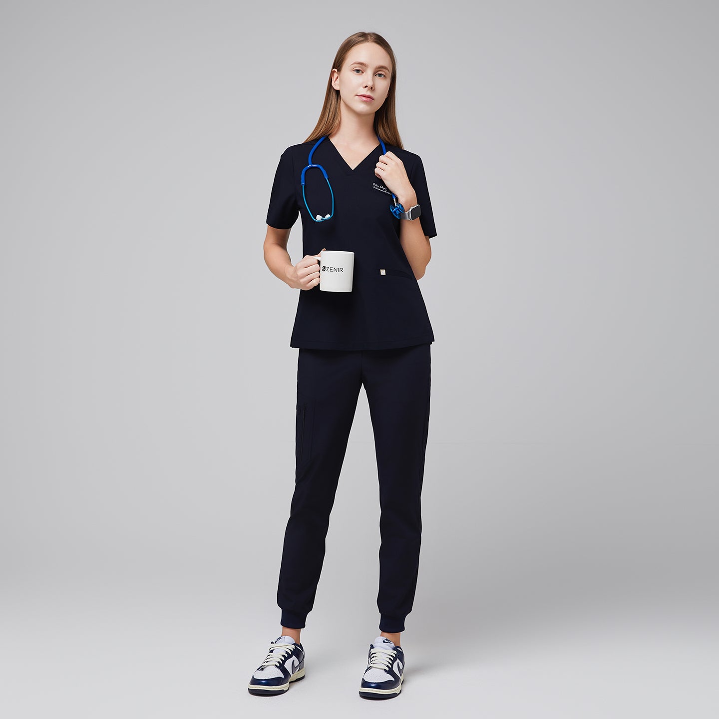 Model in an eco navy double pocket scrub top with a blue stethoscope, holding a mug, and wearing matching jogger scrub pants and sneakers,Eco Navy