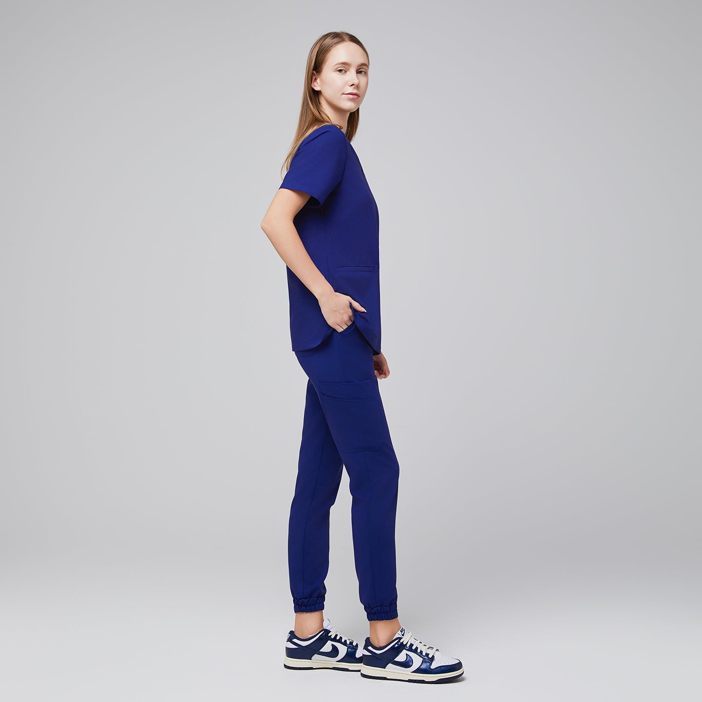 Woman in navy blue V-neck scrub top with two pockets, matching jogger scrub pants, and blue-white sneaker.Admiral Blue