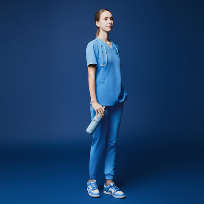 Female model in aqua blue soft stretch scrub top and pants, holding a branded water bottle and wearing a stethoscope, shown in a full-body side view against a dark blue background,Aqua Blue