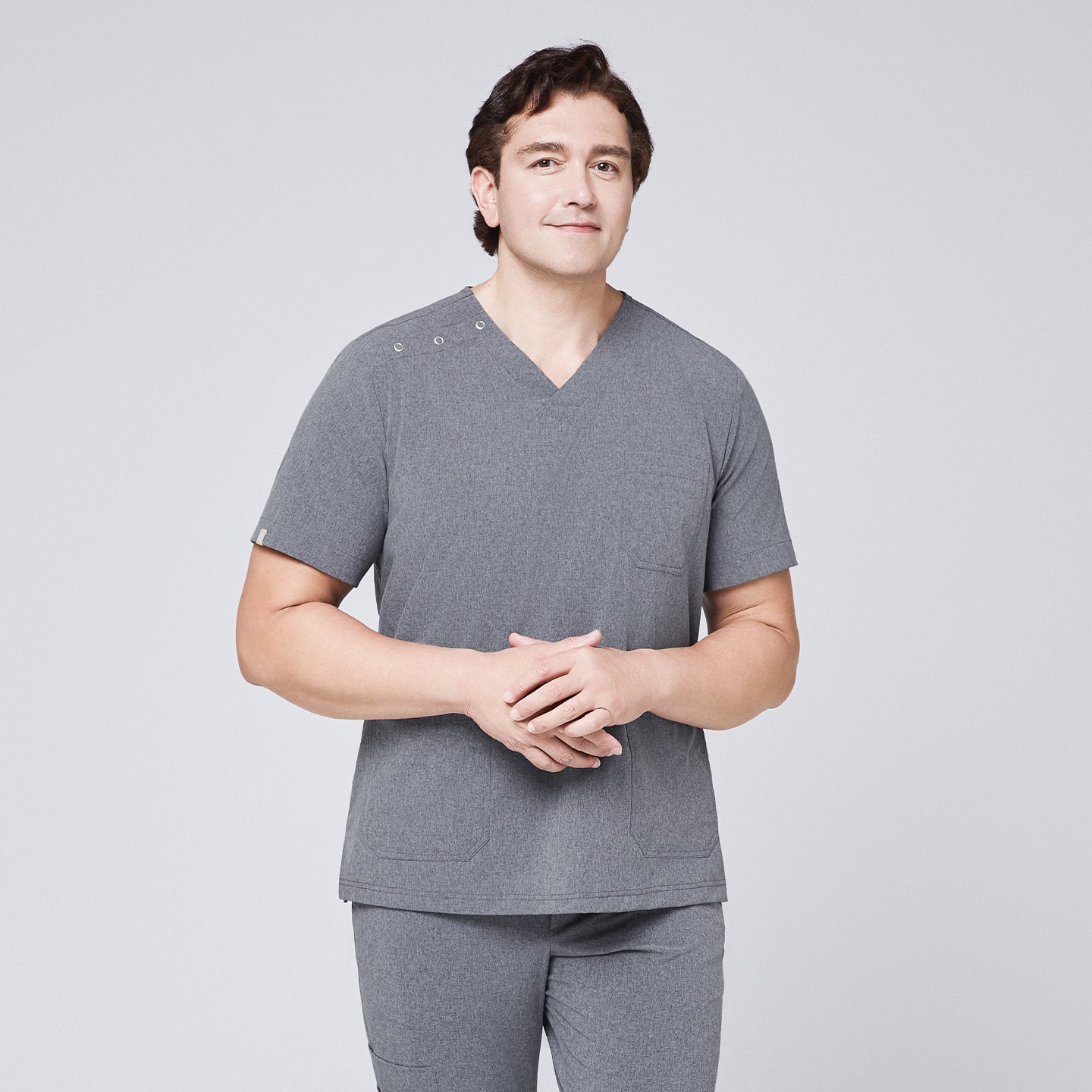 Male model wearing an ash gray scrub top with shoulder buttons and matching pants, hands clasped and smiling,Ash Gray