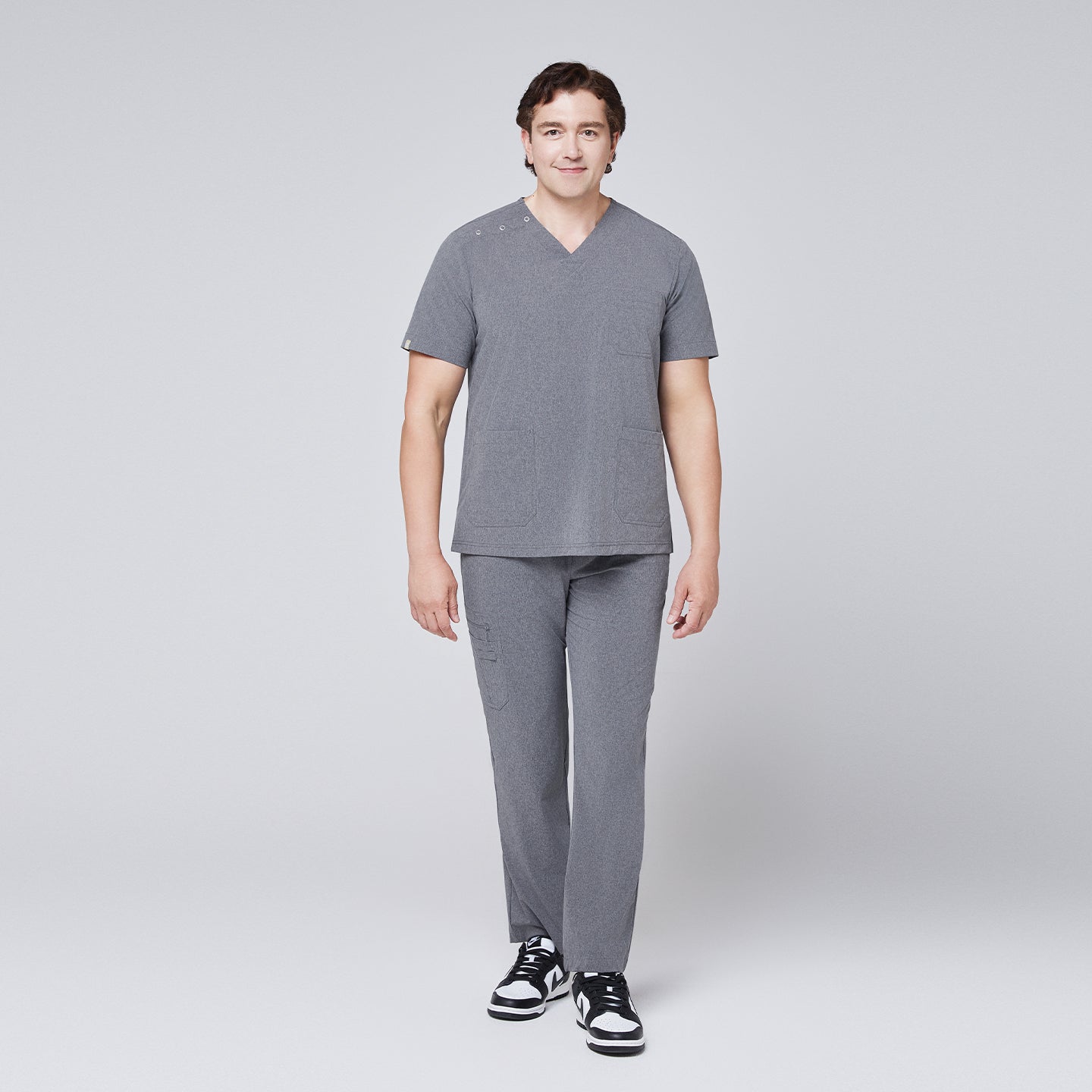 Male model in ash gray scrub set with shoulder buttons, standing and smiling in black and white sneakers,Ash Gray