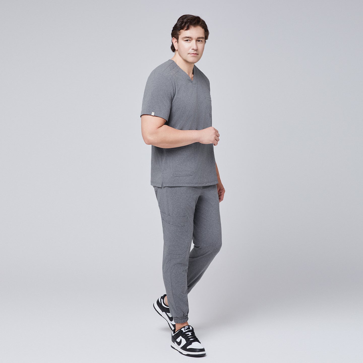 Male model in ash gray scrub top with shoulder buttons and jogger-style pants, posing in black and white sneakers,Ash Gray