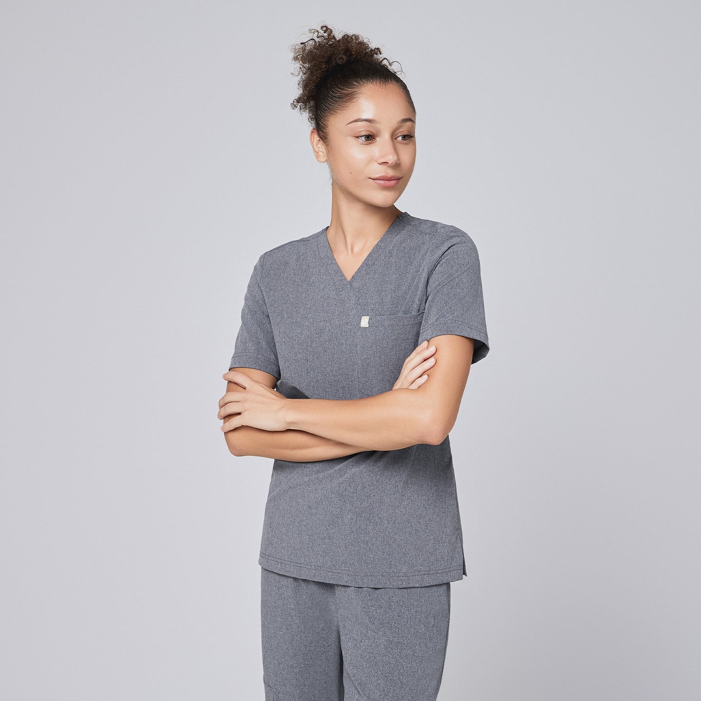 Female healthcare professional in an ash gray scrub set, standing in a rear-side pose with hands in pockets and wearing black sneakers,Ash Gray