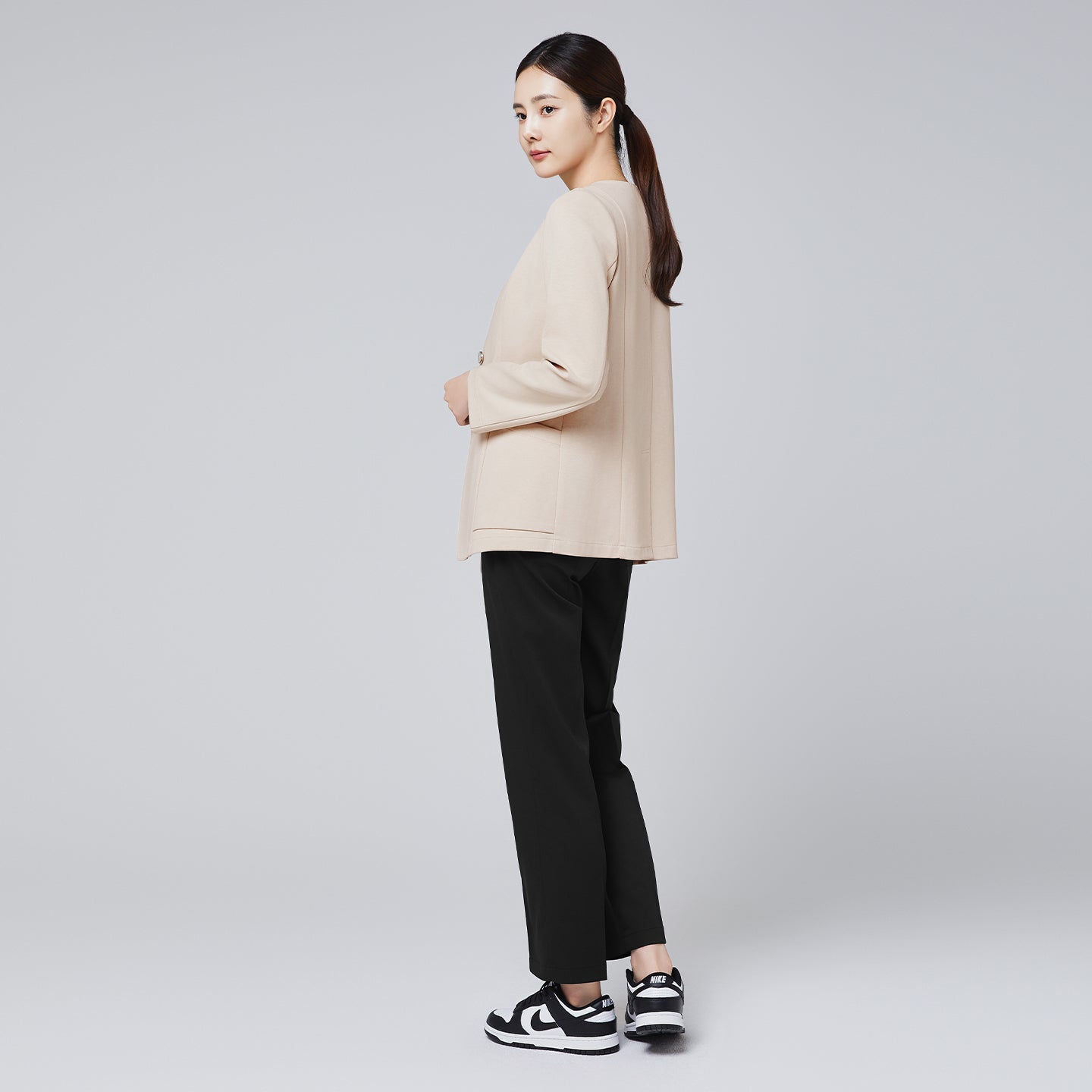 A woman wearing a beige comfort cardigan and black trousers, posing in a rear three-quarter view with her head turned toward the camera, styled with sleek sneakers and a ponytail against a plain background,Beige