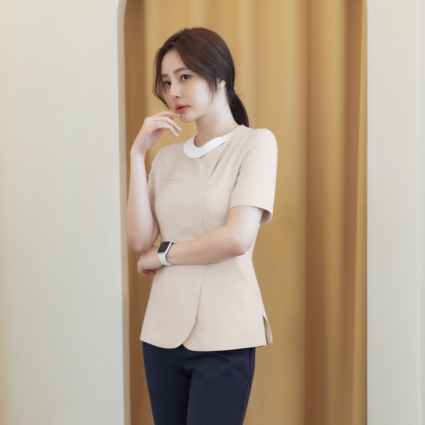 A model wearing a beige round-neck top with a front zipper, paired with dark pants, standing against a light beige and curtain background, looking thoughtful with her hand on her chin,Beige
