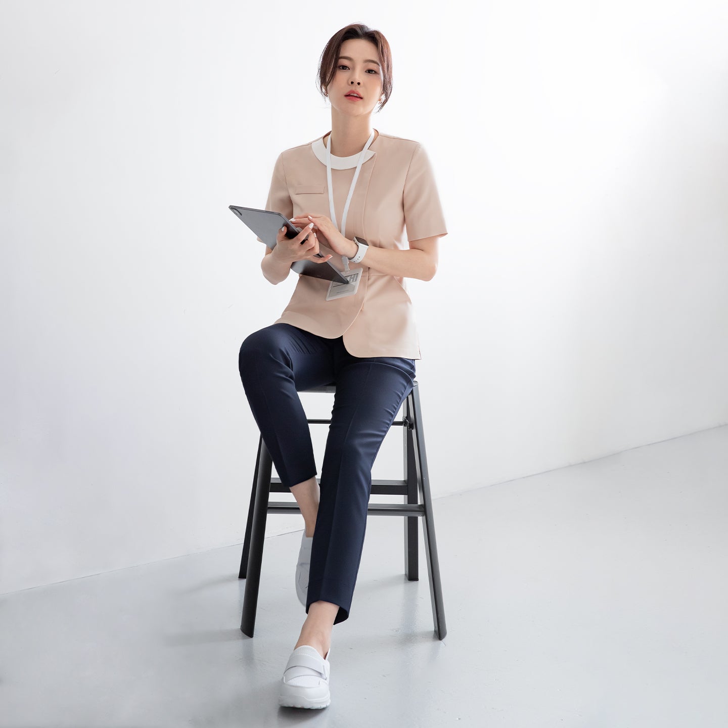 A model sitting on a stool, wearing a beige round-neck top with a front zipper, paired with dark pants and white shoes, holding a tablet and lanyard in a brightly lit room,Beige