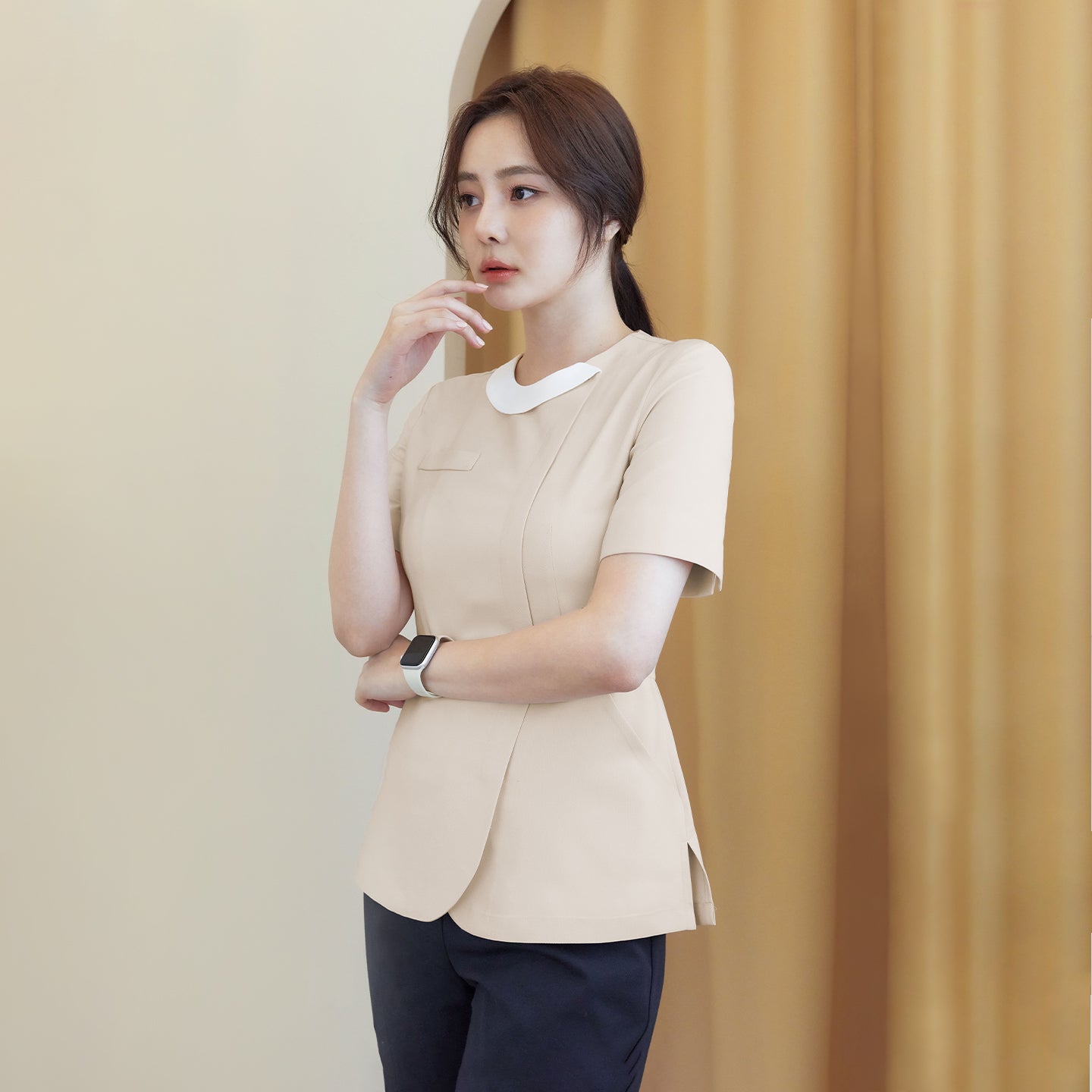 A model wearing a beige round-neck top with a front zipper, paired with dark pants, standing against a light beige and curtain background, looking thoughtful with her hand on her chinm,Beige