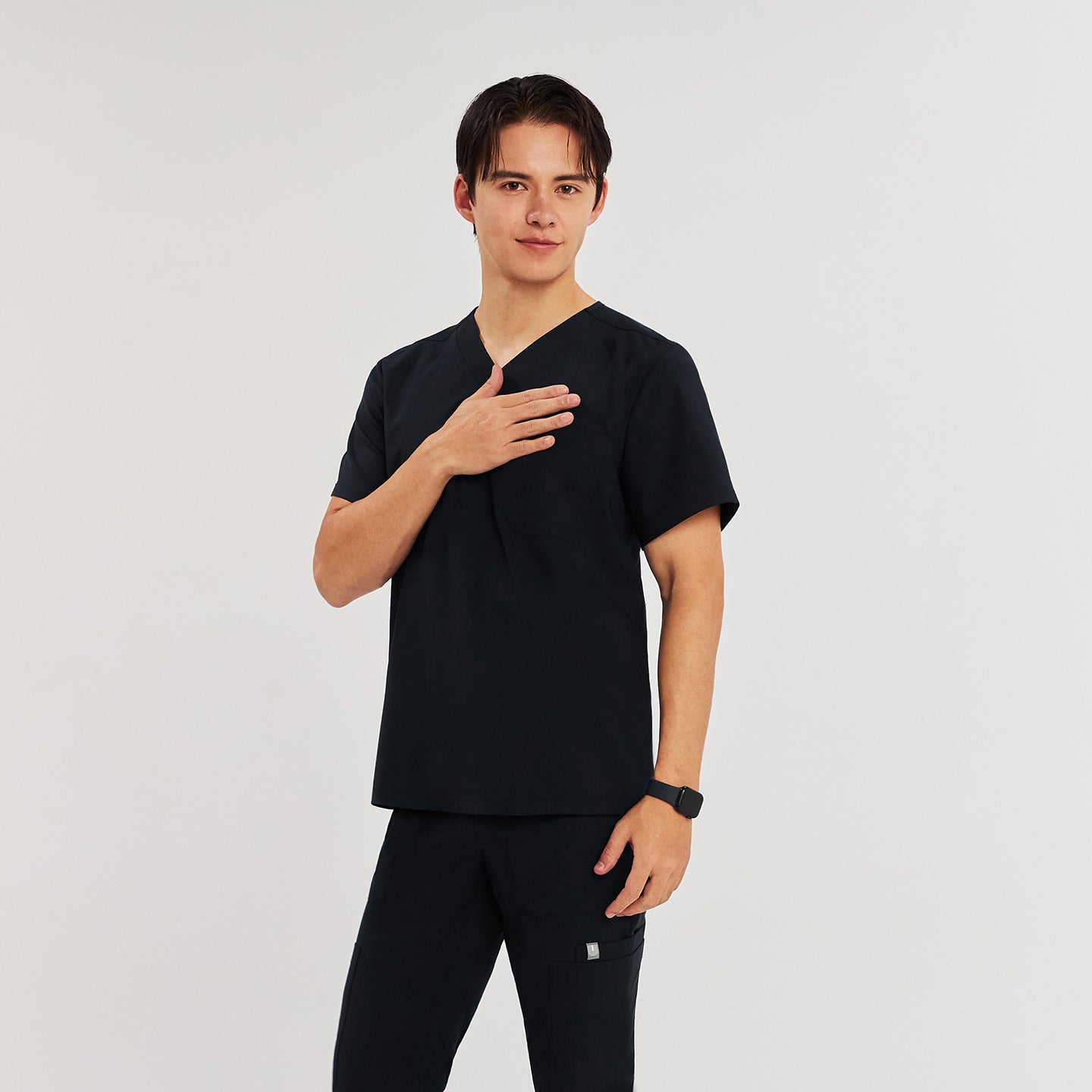 Man in black Zenir scrub top with chest pocket, standing with hand on chest, looking at the camera,Black