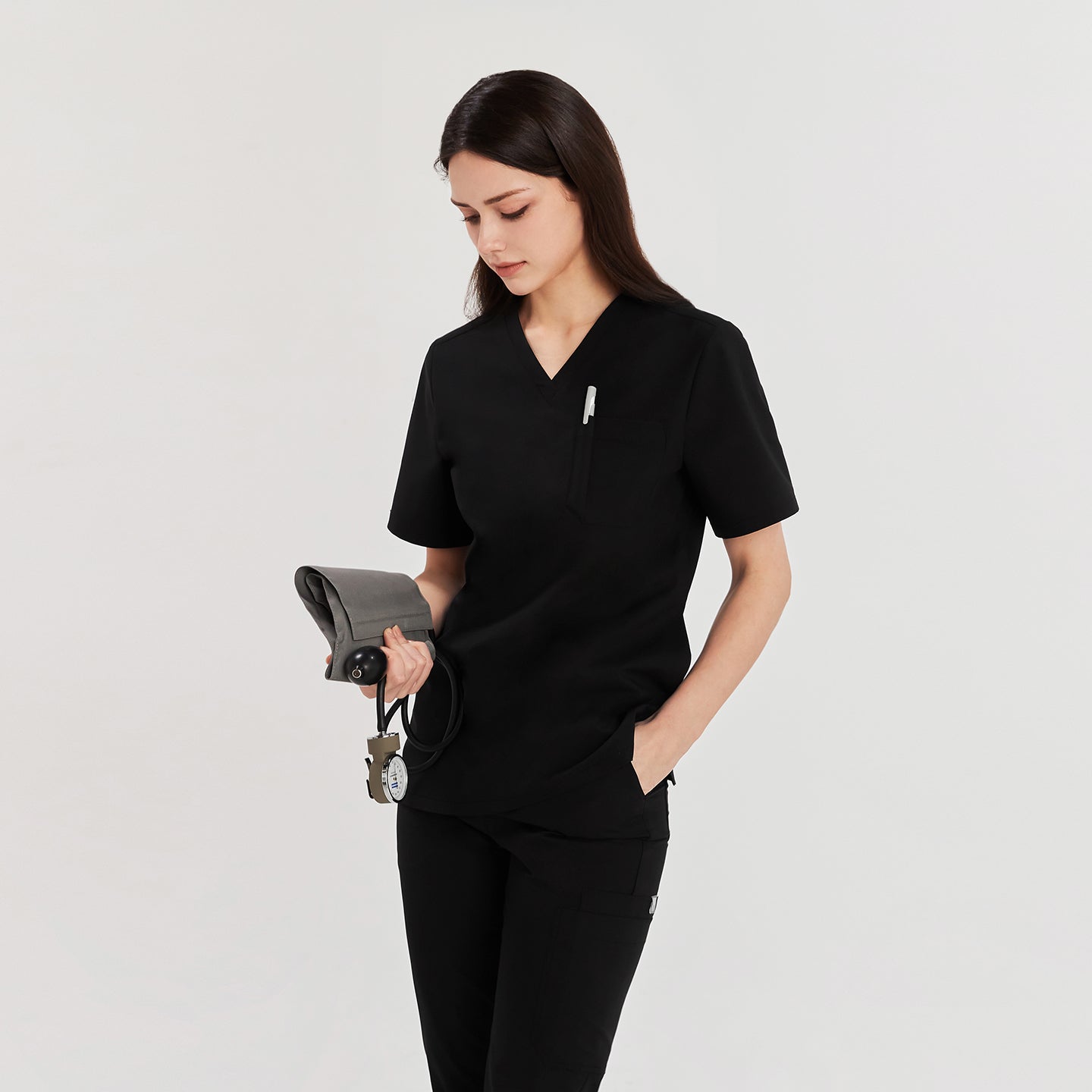 Woman in black Zenir scrub top with chest pocket, holding a blood pressure monitor, looking down, Black