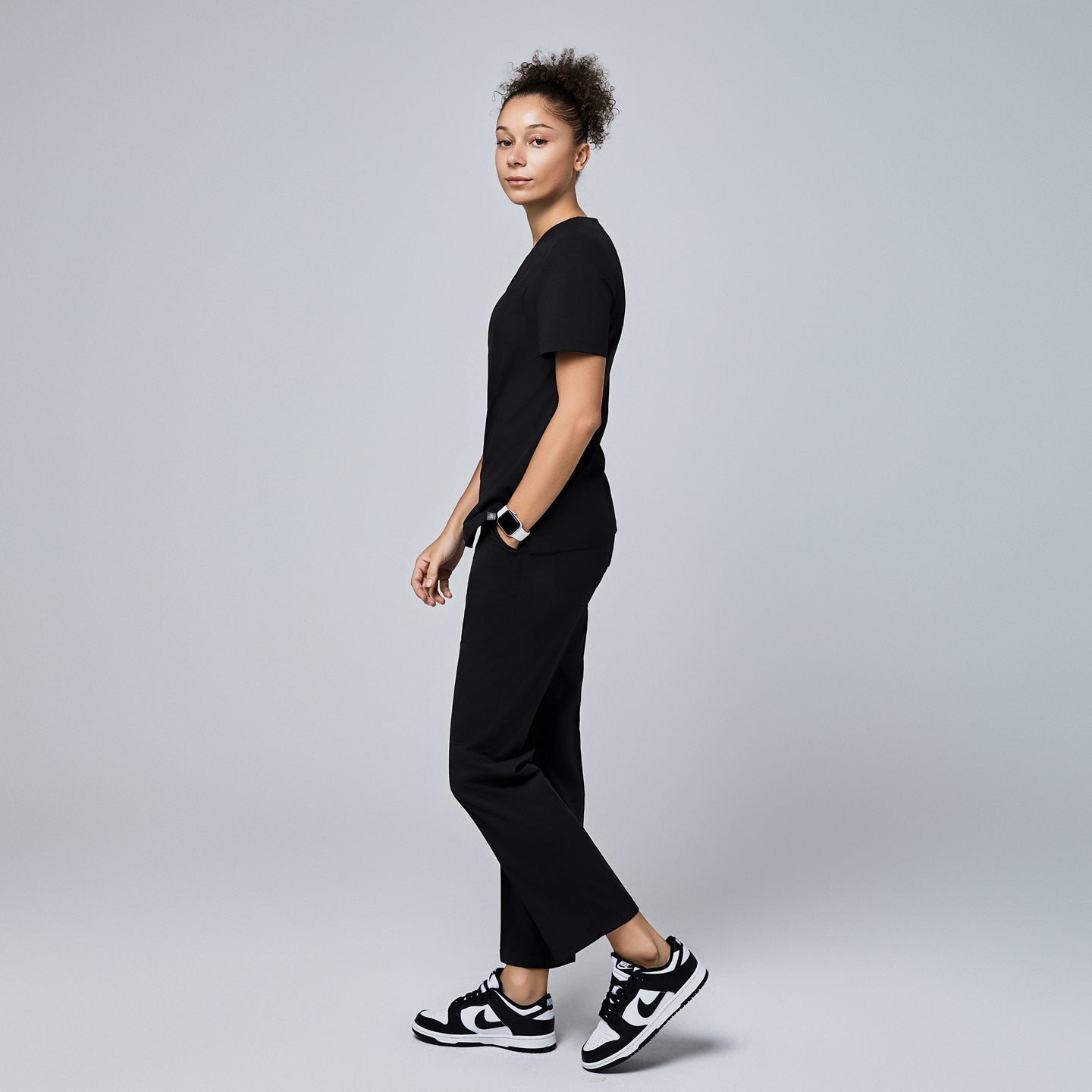 Female model wearing a black V-neck scrub top with short sleeves, paired with black scrub pants and sneakers, shown walking from a side angle,Eco Black