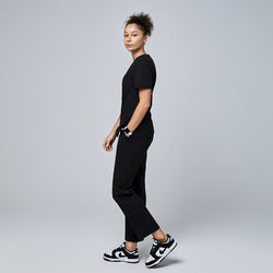 Image of Female model wearing a black V-neck scrub top with short sleeves, paired with black scrub pants and sneakers, shown walking from a side angle,Eco Black