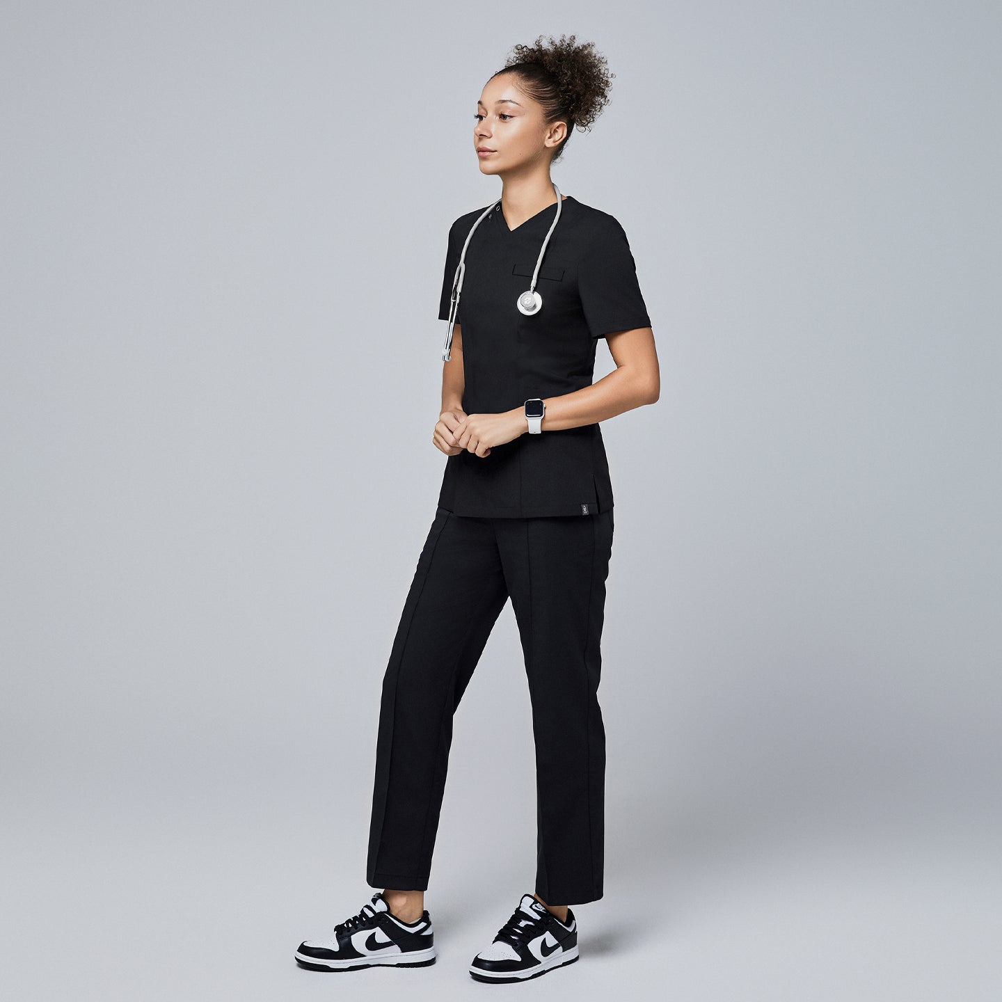 Female model wearing a black V-neck scrub top with a front zipper, chest pocket, and short sleeves, accessorized with a stethoscope and paired with matching black scrub pants and sneakers,Eco Black