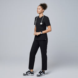 Image of Female model wearing a black V-neck scrub top with a front zipper, chest pocket, and short sleeves, accessorized with a stethoscope and paired with matching black scrub pants and sneakers,Eco Black