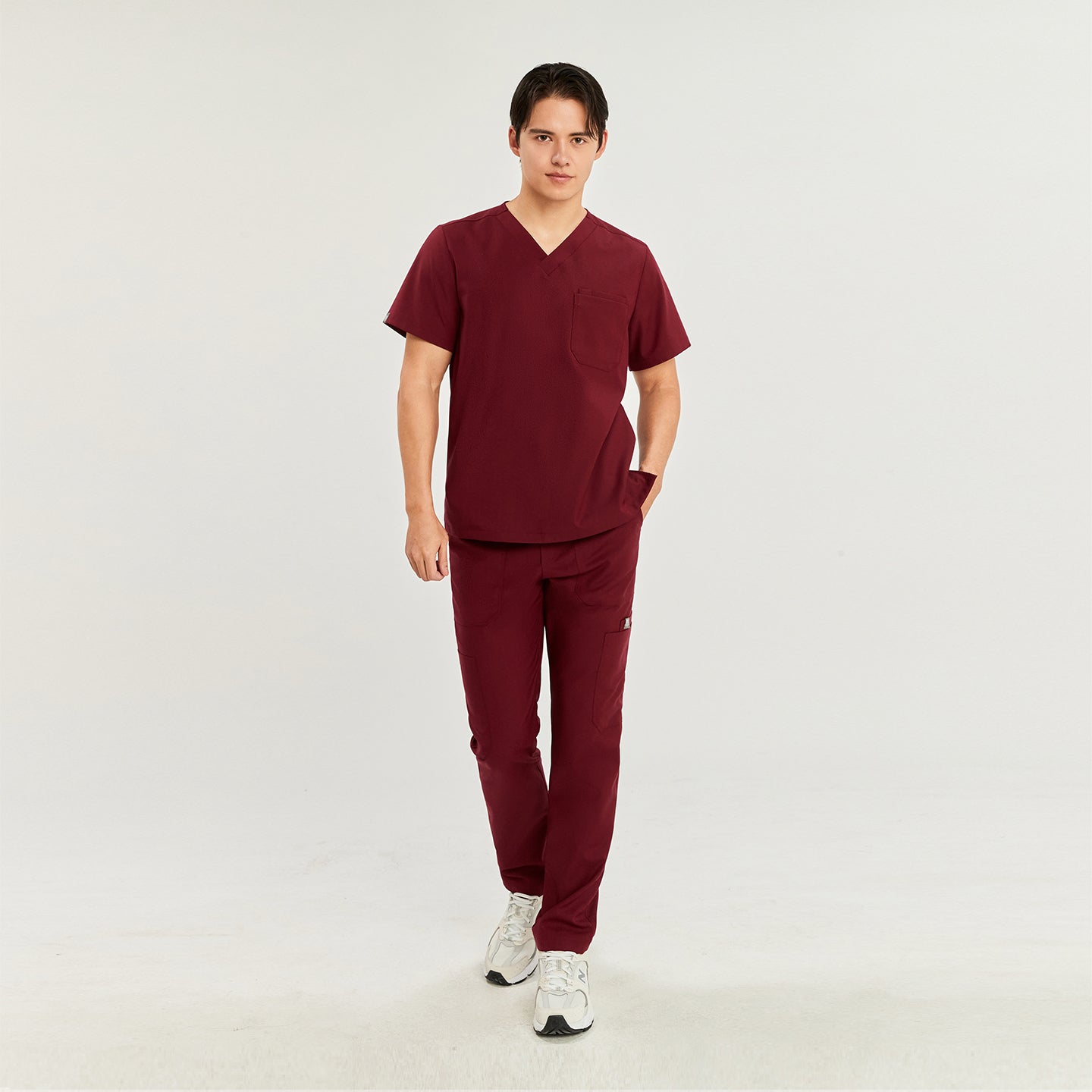 Male model wearing a scrub top with a chest pocket, paired with matching scrub pants and white sneakers, standing with one hand in his pocket,Burgundy