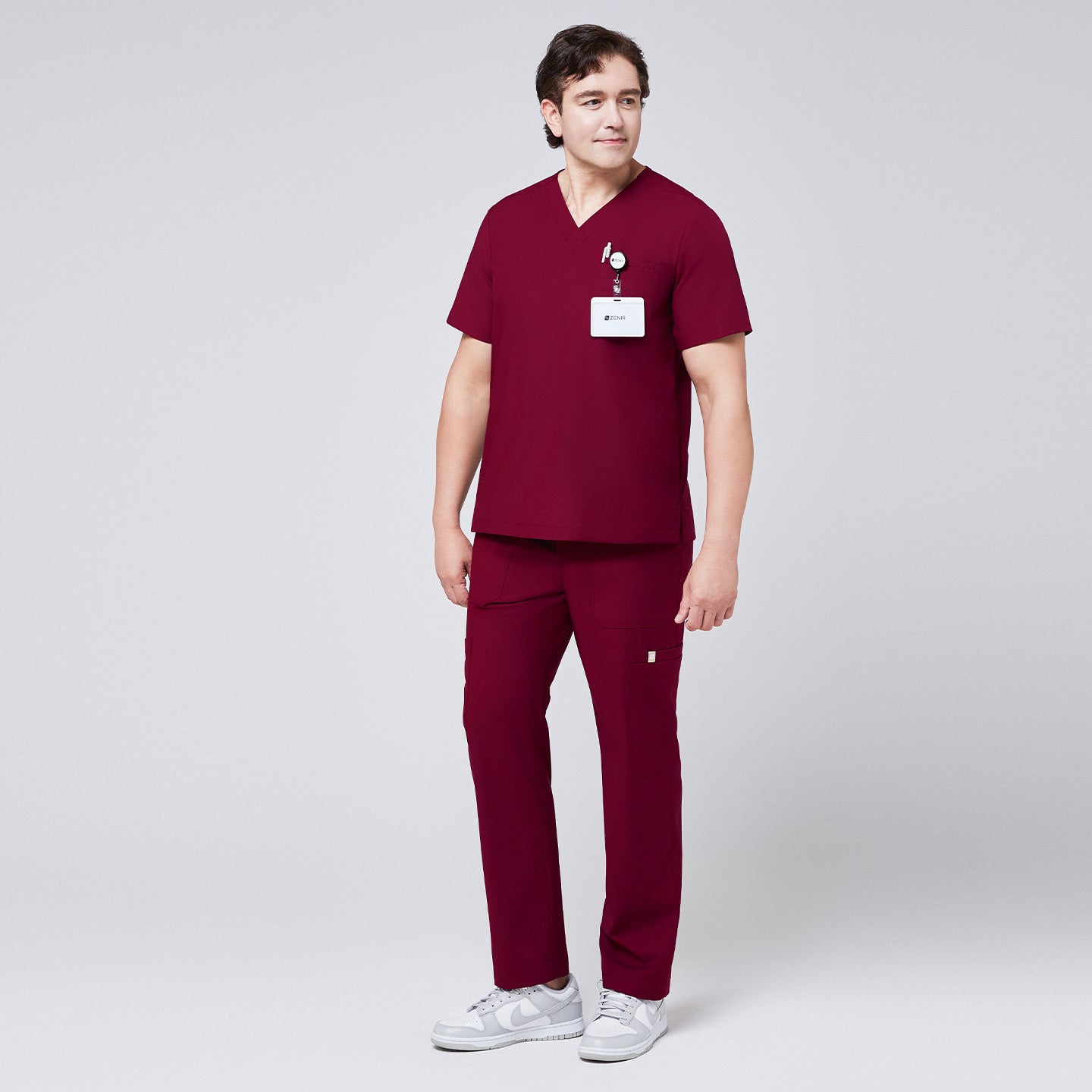 Man wearing burgundy V-neck scrub top and 8-pocket straight scrub pants, accessorized with a name badge and paired with white sneakers,Burgundy