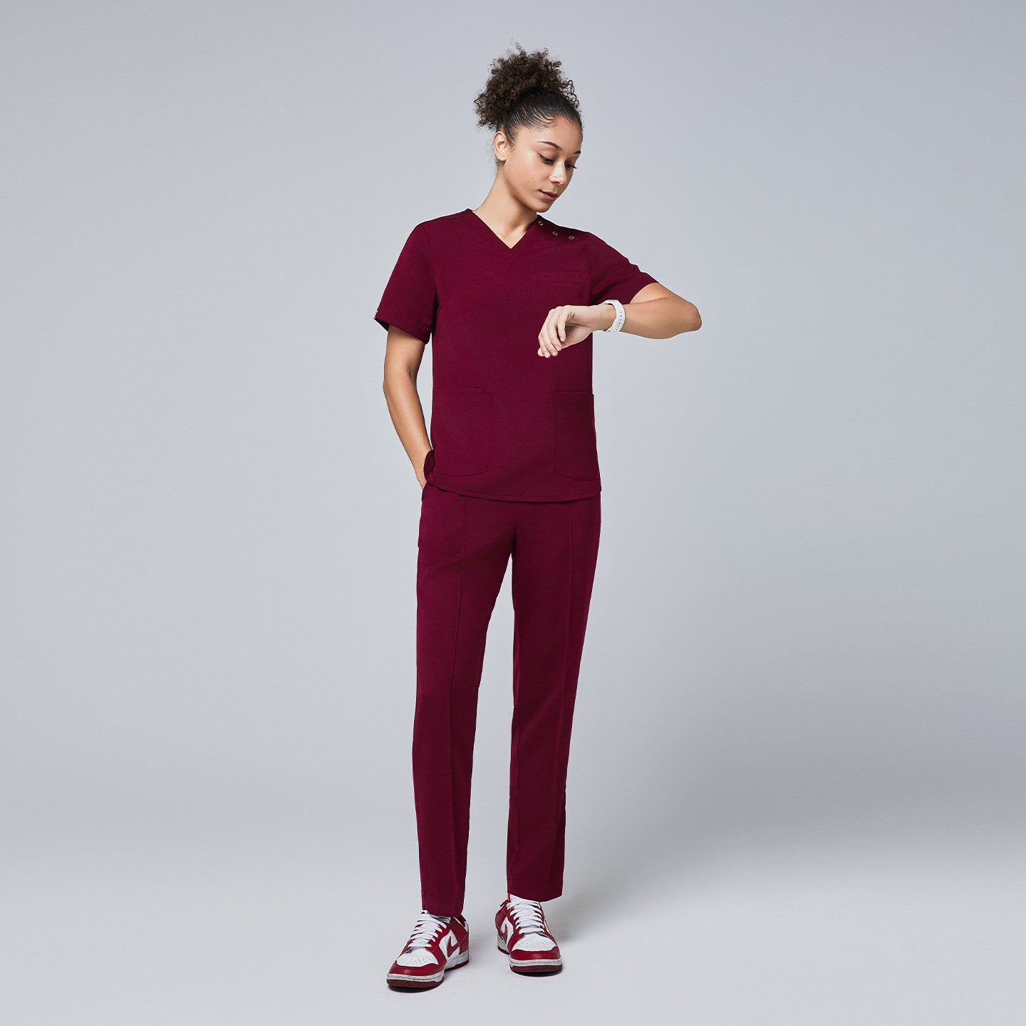 Full-body view of a woman wearing a burgundy scrub set with straight-leg pants and sneakers, checking the time on her smartwatch,Burgundy